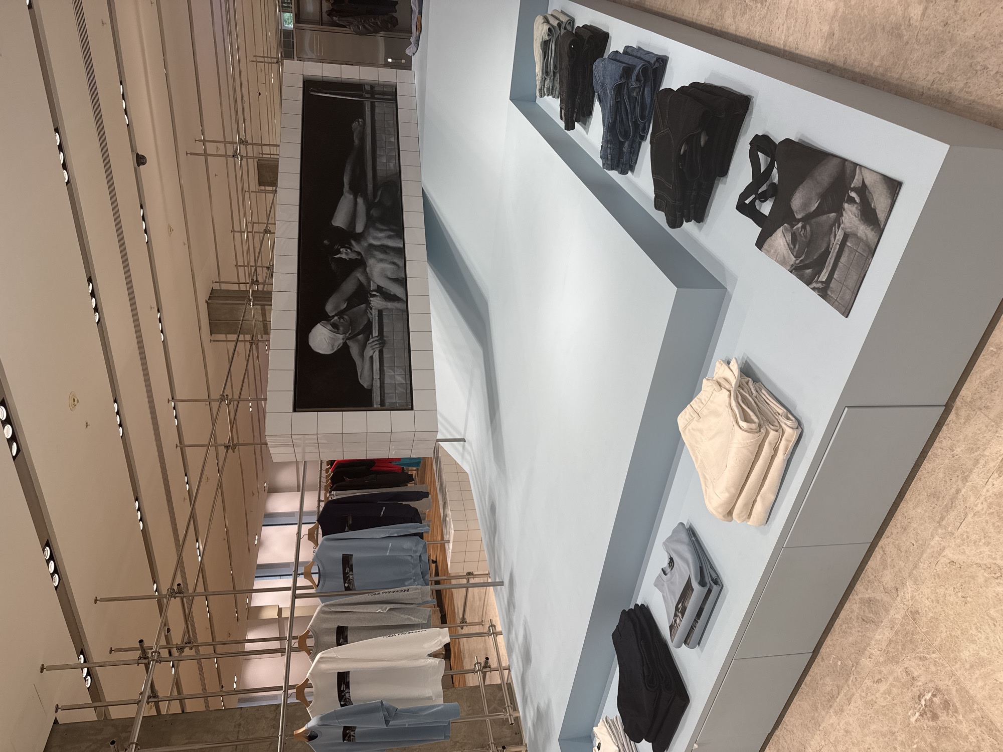 The image depicts a retail store interior, likely a clothing boutique, with a minimalist aesthetic. In the foreground, a light blue display shelf holds folded clothing items, including dark trousers, a light blue t-shirt, and a stack of beige pants. To the right, a black tote bag with a grayscale image printed on it is displayed. Further back, another shelf displays folded jeans and dark trousers. The background features a large mural or artwork depicting two people in what appears to be a swimming pool. Above this artwork, a metal scaffolding system is visible, suggesting a temporary installation or a modern design element. Clothing items, including sweatshirts and t-shirts in various colors (light blue, white, gray, and navy blue), are hung on a rack in the upper left portion of the image. The store has a light-colored tiled floor and a white ceiling with recessed lighting. The overall impression is one of a well-organized and visually curated retail space. The location is identified as a store in Moscow, Russia, based on the context provided.