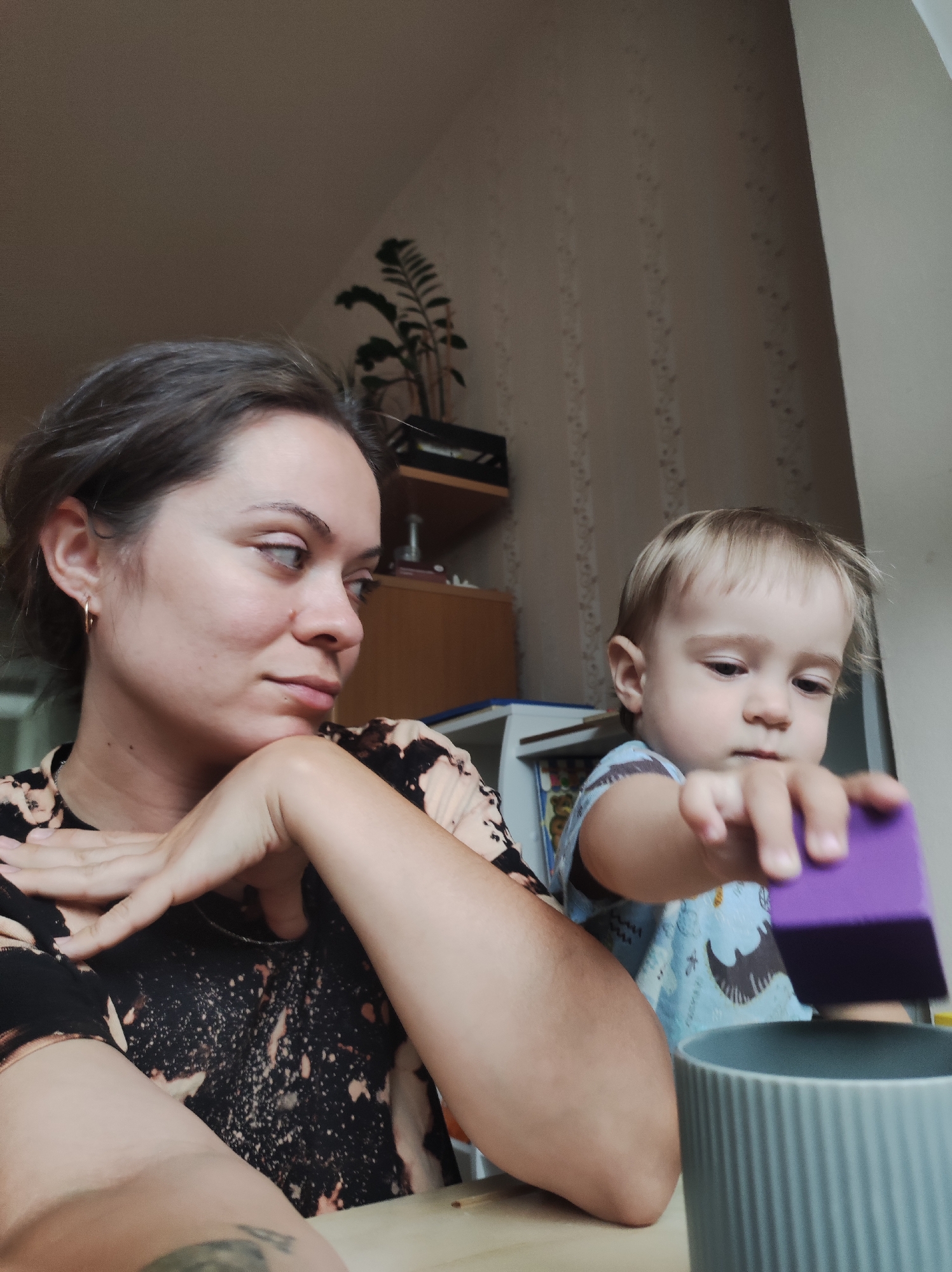 A woman and a toddler are indoors. The woman, with her dark hair pulled back, rests her chin on her hand and gazes slightly away with a pensive expression. She wears a black and beige patterned top. A young child, with blonde hair, stands next to her, holding a purple block over a gray ribbed container. The location is Chaikovsky, Russia, as determined from metadata associated with this photograph.