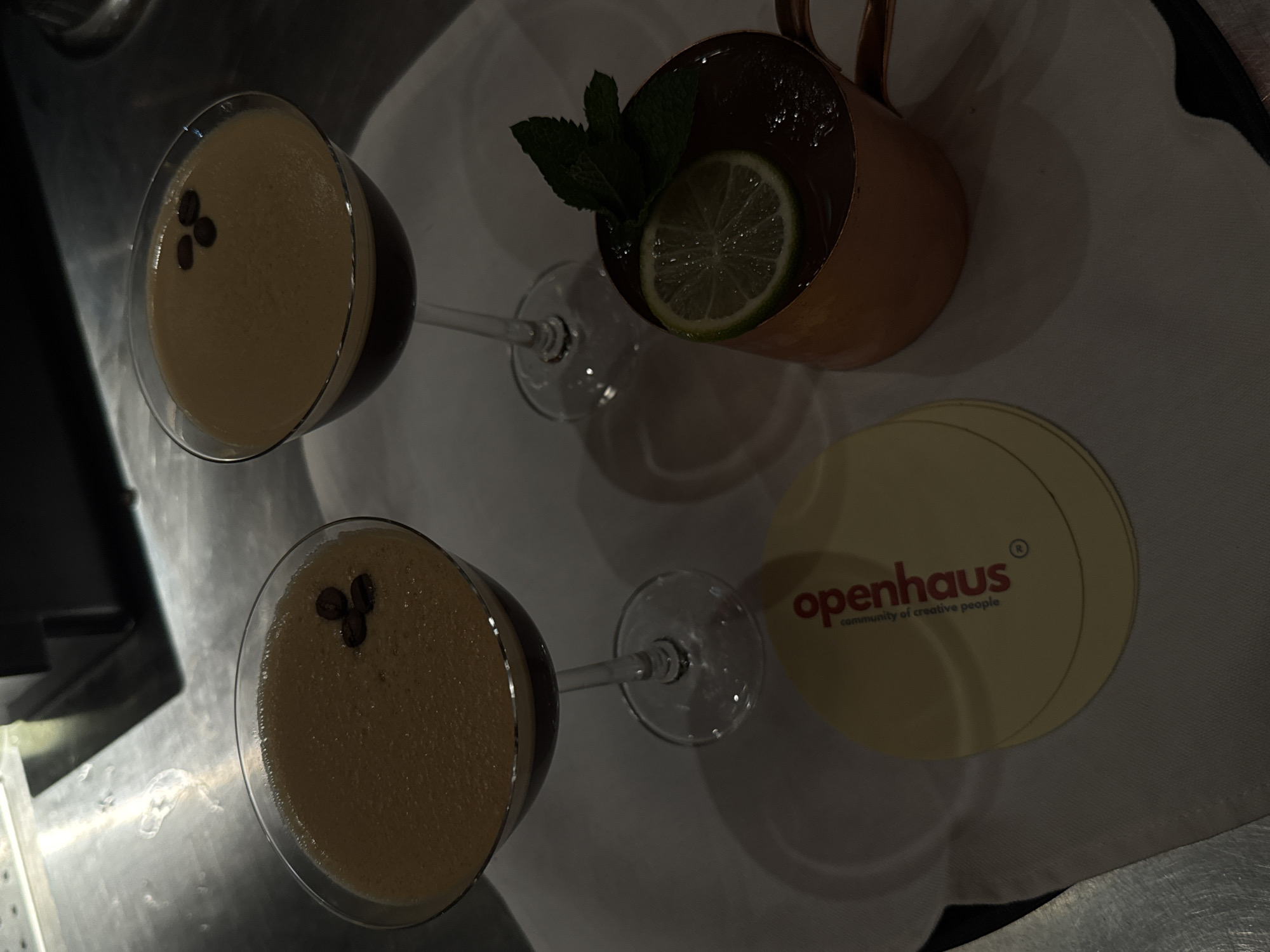 The image shows a top-down view of two espresso martinis in stemmed cocktail glasses, each garnished with three coffee beans. Next to them is a copper mug containing a drink garnished with a lime wedge and mint leaves. Adjacent to the mug is a round coaster with the "openhaus community of creative people" logo in red text. The scene appears to be indoors, possibly a bar or restaurant setting, with a metallic surface visible. The location in Yerevan, Armenia, is not identifiable from the visual information provided.