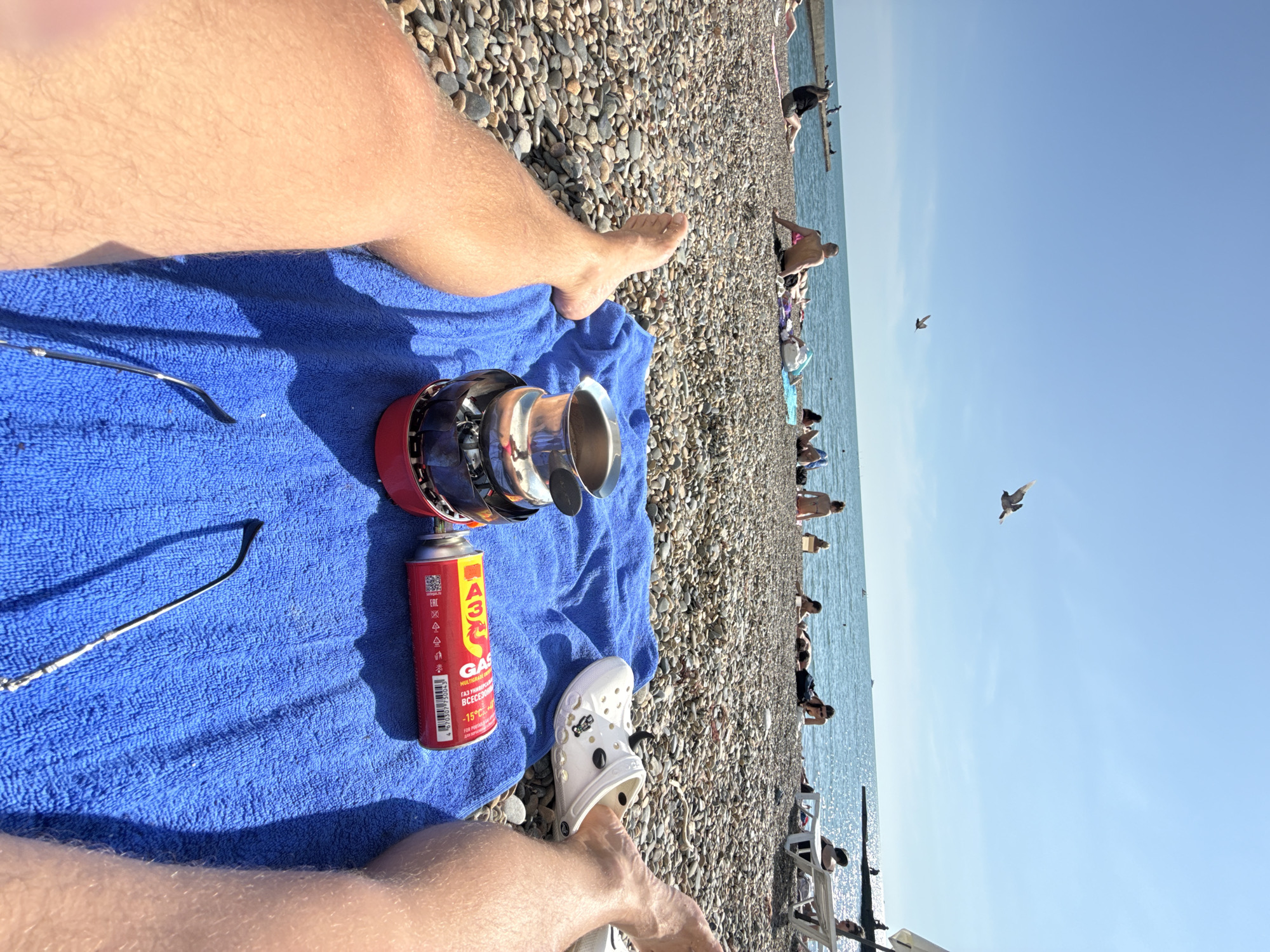 This is a first-person perspective shot of a person relaxing on a pebble beach in Sochi, Russia. The person's legs are in the foreground, with a blue towel laid out on the pebbles. On the towel are a portable camping stove with a metal cup on top, a can of gas, a pair of sunglasses, and a white Croc shoe. In the background, several people are sunbathing and sitting on the beach, with lounge chairs and umbrellas visible. The Black Sea is calm and sparkling under a clear blue sky, with a few boats in the distance. A bird is flying in the sky. The overall atmosphere is one of leisure and enjoyment of a sunny day by the sea.