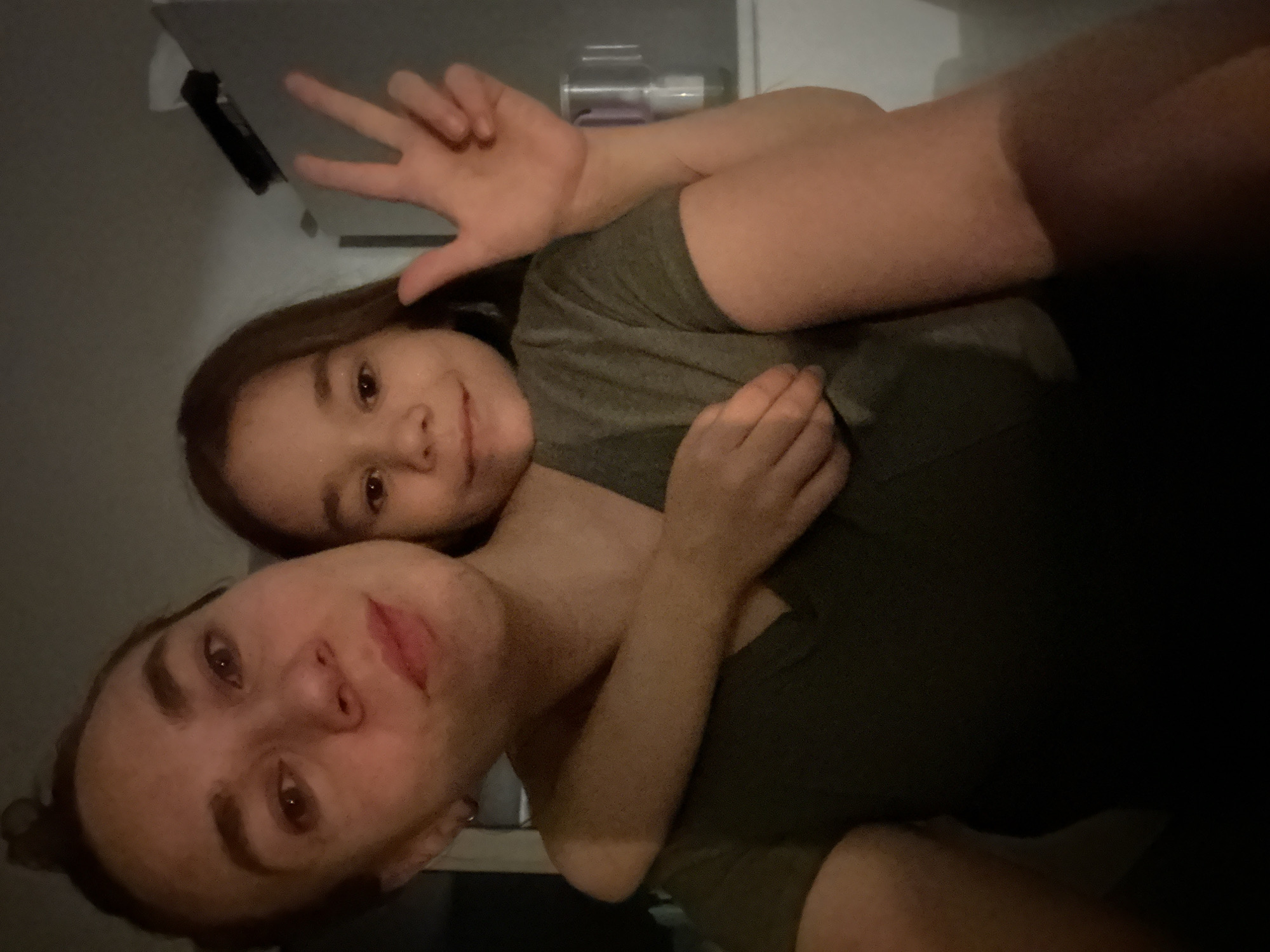 A selfie featuring an adult woman and a young girl. The girl is positioned behind the woman, with her arms wrapped around the woman's neck. The girl is displaying a peace sign with her fingers. Both are wearing similarly colored olive green t-shirts. The background includes a partially visible cabinet and indistinct items. The location is unspecified and unidentifiable from the image. The scene is indoor. There is no evidence to indicate it being in the city of Kharnavy, Russia.