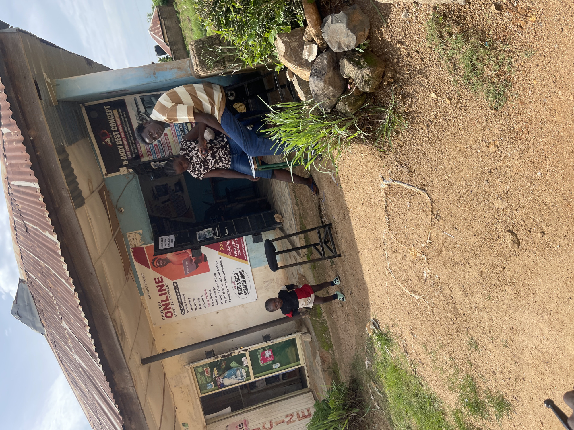 A wide shot captures a scene in front of a small shop in Rafin Par, Nigeria. The shop, constructed with a corrugated metal roof and a simple facade, displays several large posters advertising services. One prominent poster announces "GENERAL ONLINE SERVICES & PAYMENT" and lists various printing and computer-related services. Another poster, to the right, features "D-ANDY BEST CONCEPT" and "COMPUTER TRAINING INSTITUTE."
In the foreground, a young child, appearing to be a toddler, stands facing the camera, dressed in a red, black, and white outfit. To the right of the child, a black metal stool with a round seat is placed on the dusty ground.
In the background, a man and a woman are seated near the shop. The man, wearing a striped t-shirt and blue jeans, is looking towards the camera. The woman, in a patterned dress and blue shorts, is looking down at something on her lap. They appear to be interacting or resting in front of the establishment.
The environment is characterized by a dusty, reddish-brown ground, sparse vegetation, and a few rocks scattered around. The sky above is partly cloudy. The overall impression is a snapshot of everyday life in a commercial area in Rafin Par.