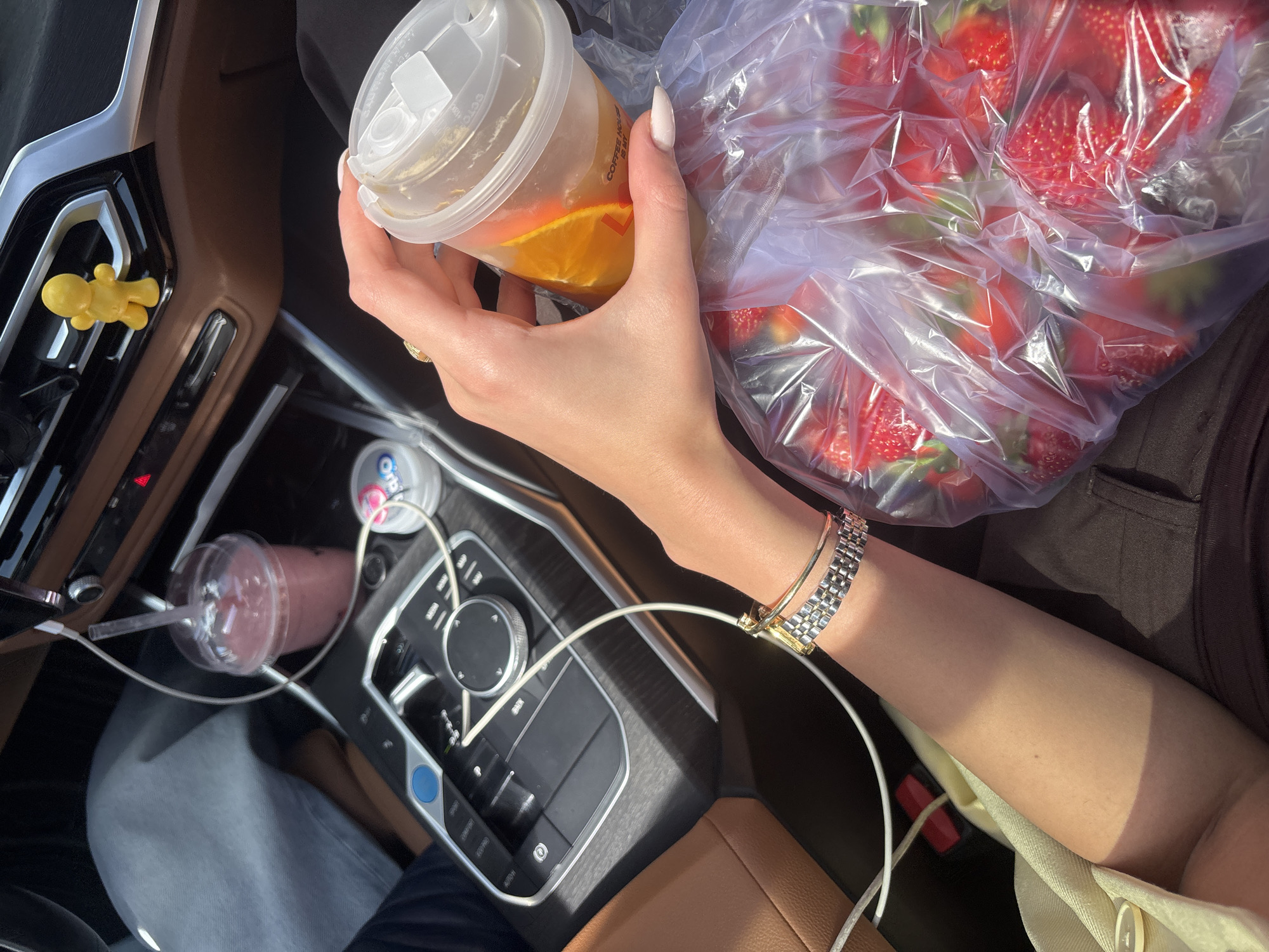 Inside a car, an individual holds a plastic cup containing orange slices and liquid, alongside a clear plastic bag filled with strawberries. A pink drink in a cup with a straw is visible in the center console. The car's interior features brown leather seats and a modern dashboard with electronic controls. A yellow figure is attached to the air vent. The location is potentially Yerevan, Armenia.