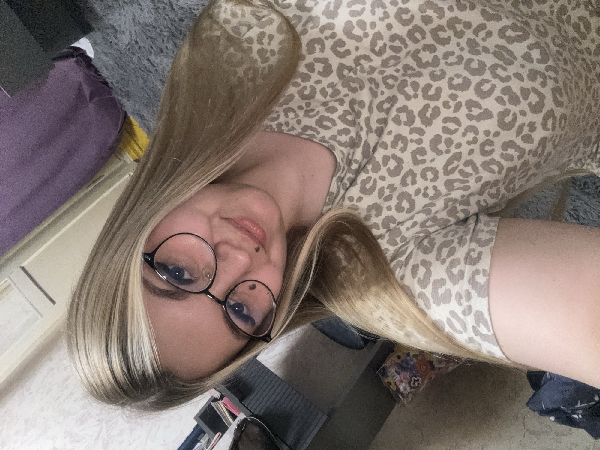 The image is a close-up, selfie-style photograph of a young woman with blonde hair and glasses, wearing a light-colored t-shirt with a leopard print pattern. She is looking towards the camera with a slight smile. The background appears to be an indoor domestic setting, possibly a bedroom, with a gray fuzzy rug, a dark gray furniture piece on the left, and what looks like a purple blanket or curtain in the upper right corner. The lighting suggests an indoor environment, possibly with artificial light. There is no indication of the location being in the city of Большой Букор, Russia.