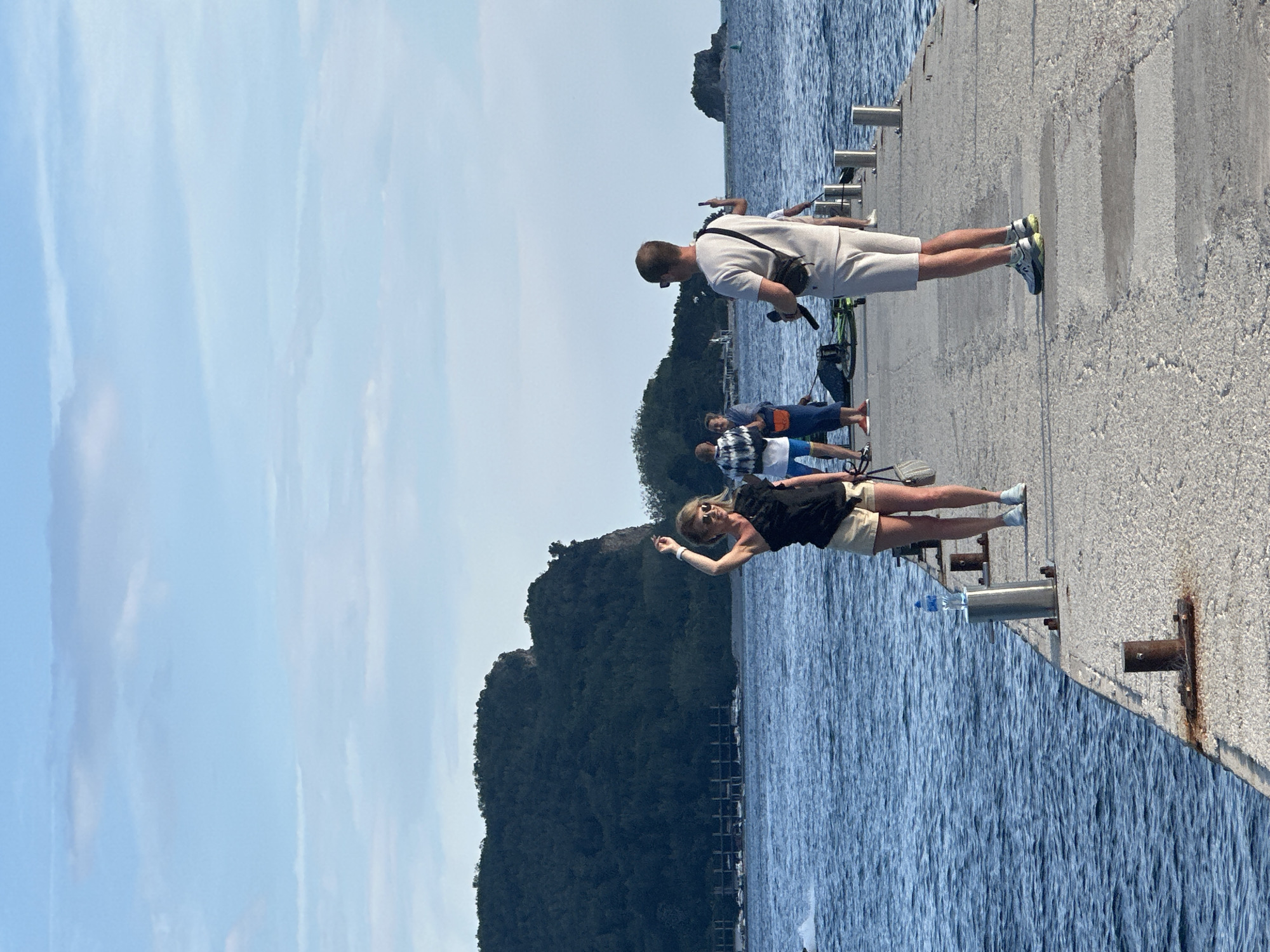 On a sunny day in Budva, Montenegro, a concrete pier juts out into the blue waters of the Adriatic Sea. A group of people are on the pier: a woman in a black top and tan shorts raises her arm, a man in a white outfit looks down at the pier, and other individuals stand further back. A bottle of water sits on the edge of the pier. In the background, a tree-covered hill rises from the water. The sky is partly cloudy.