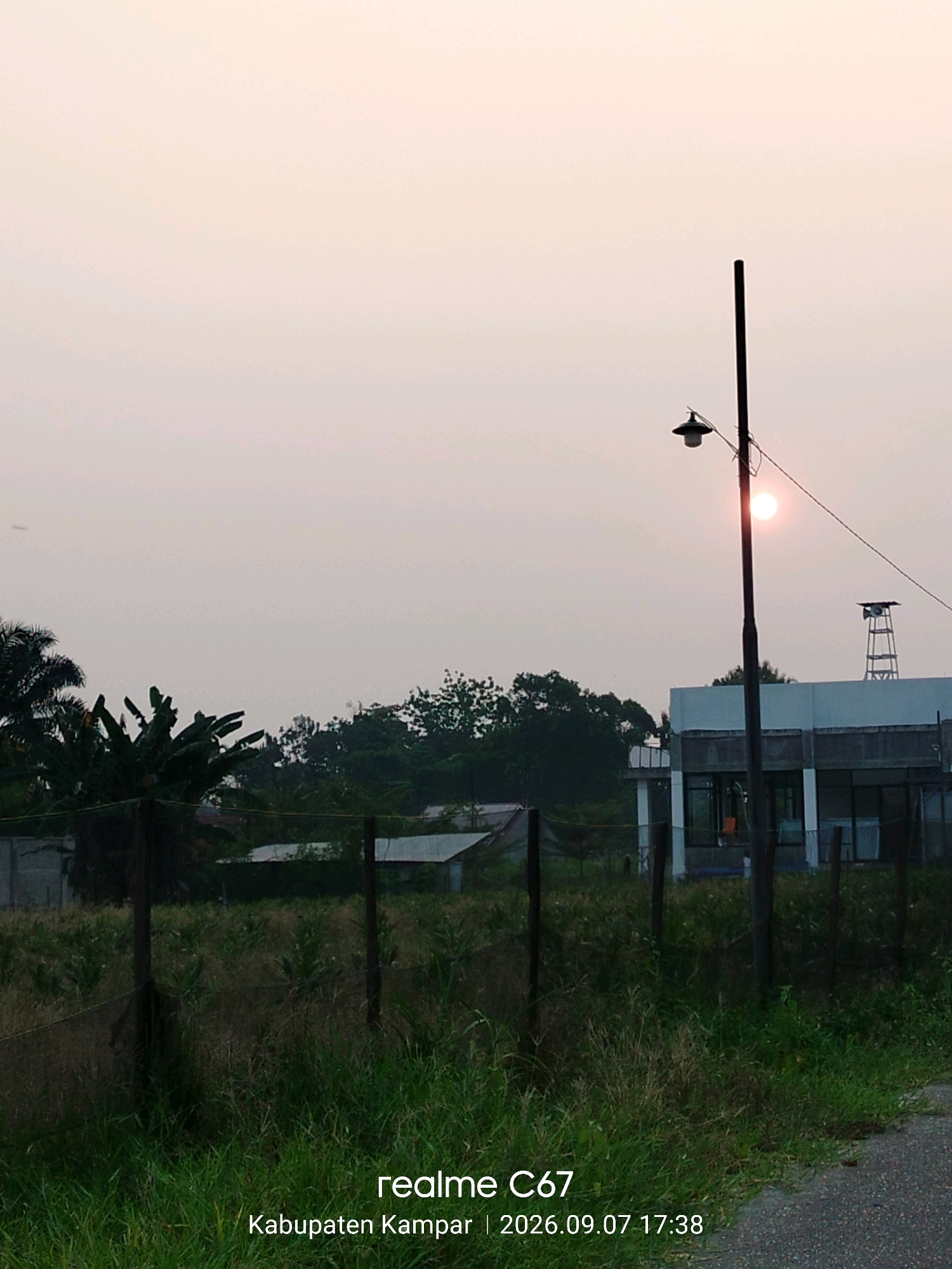 This is a landscape photo captured at dusk in Pandau Jaya, Indonesia. The sun, appearing as a small, bright orange orb, is low in the sky, suggesting the photo was taken during sunset. The sky is hazy and pale, creating a soft, diffused light.
The scene depicts an outdoor setting with a mix of natural and man-made elements. In the foreground, tall, dry grass and some green vegetation are visible, bordered by a fence made of posts and mesh wire. A paved surface, possibly a road or pathway, is present on the right side.
Dominating the mid-ground is a modern building with large windows and a white facade. To the left of the building, a dark lamppost with a distinct lantern stands prominently. Behind the building and to the left, a cluster of trees, including palm trees, creates a dense green backdrop. A smaller, shed-like structure is partially visible behind the fence. A tall, skeletal tower with what appears to be loudspeakers is situated to the right of the main building.
The overall mood is serene and somewhat quiet, characteristic of the twilight hours. There is no indication of human activity or emotional expression.
Visible text overlays on the image read: "realme C67" and "Kabupaten Kampar | 2026.09.07 17:38". The date suggests this photo was taken in the future.