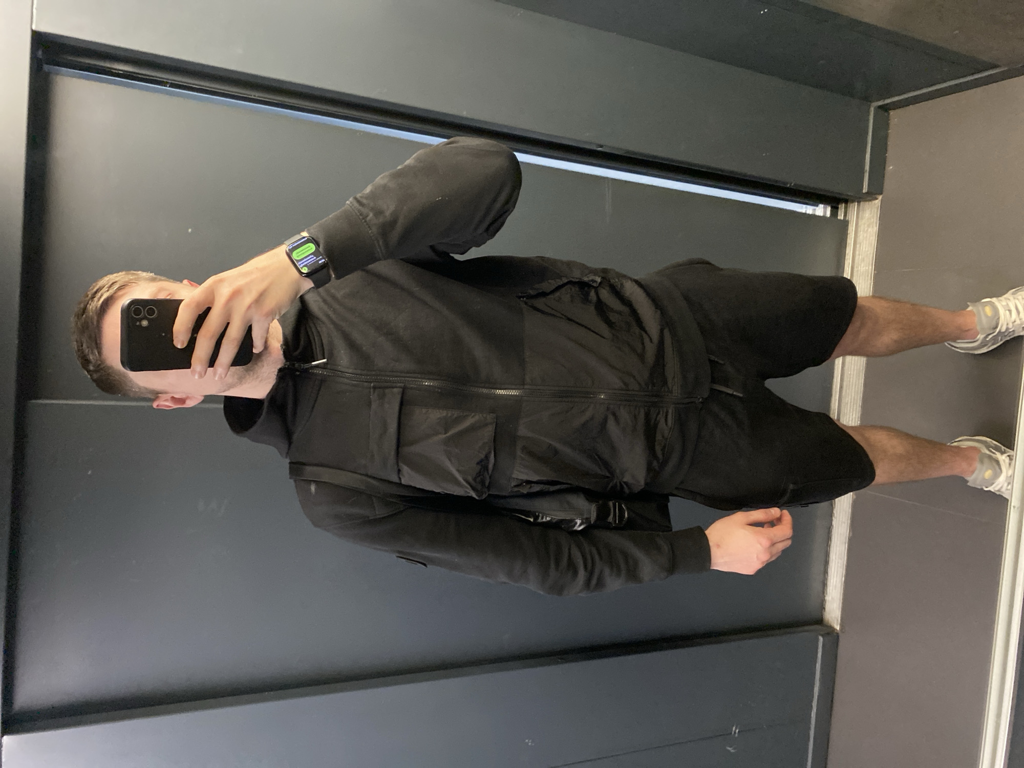 In Moscow, Russia, a young man takes a mirror selfie in an elevator. He is wearing a black hooded jacket with a zippered front and two chest pockets, black shorts, and grey and white sneakers. He is also wearing a black smartwatch on his left wrist and is holding a black smartphone in his right hand, obscuring his face. A black backpack is slung over his left shoulder. The elevator walls are dark grey, with a metallic strip along the bottom of the doors. The lighting is neutral.