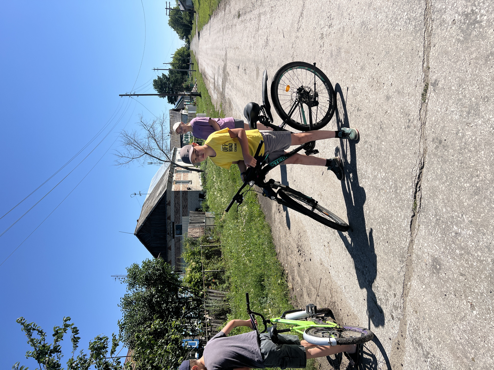 In Pivkino, Russia, on a bright, sunny day, two boys and a girl stand on a dirt road lined with houses and vegetation. The boy in the foreground, wearing a yellow t-shirt with "OFF ROAD EXTREME" printed on it, stands next to a black bicycle with a green accent on the rim. He wears a grey baseball cap and dark sneakers. To his left, another boy, partially visible from the back, is dressed in a grey t-shirt and shorts, standing with a lime green bicycle that has a purple rim. Behind them, a girl in a purple t-shirt and a white baseball cap looks towards the right side of the frame. The road stretches into the distance, with houses on either side and power lines overhead. The environment appears rural, with unpaved roads and a clear blue sky.