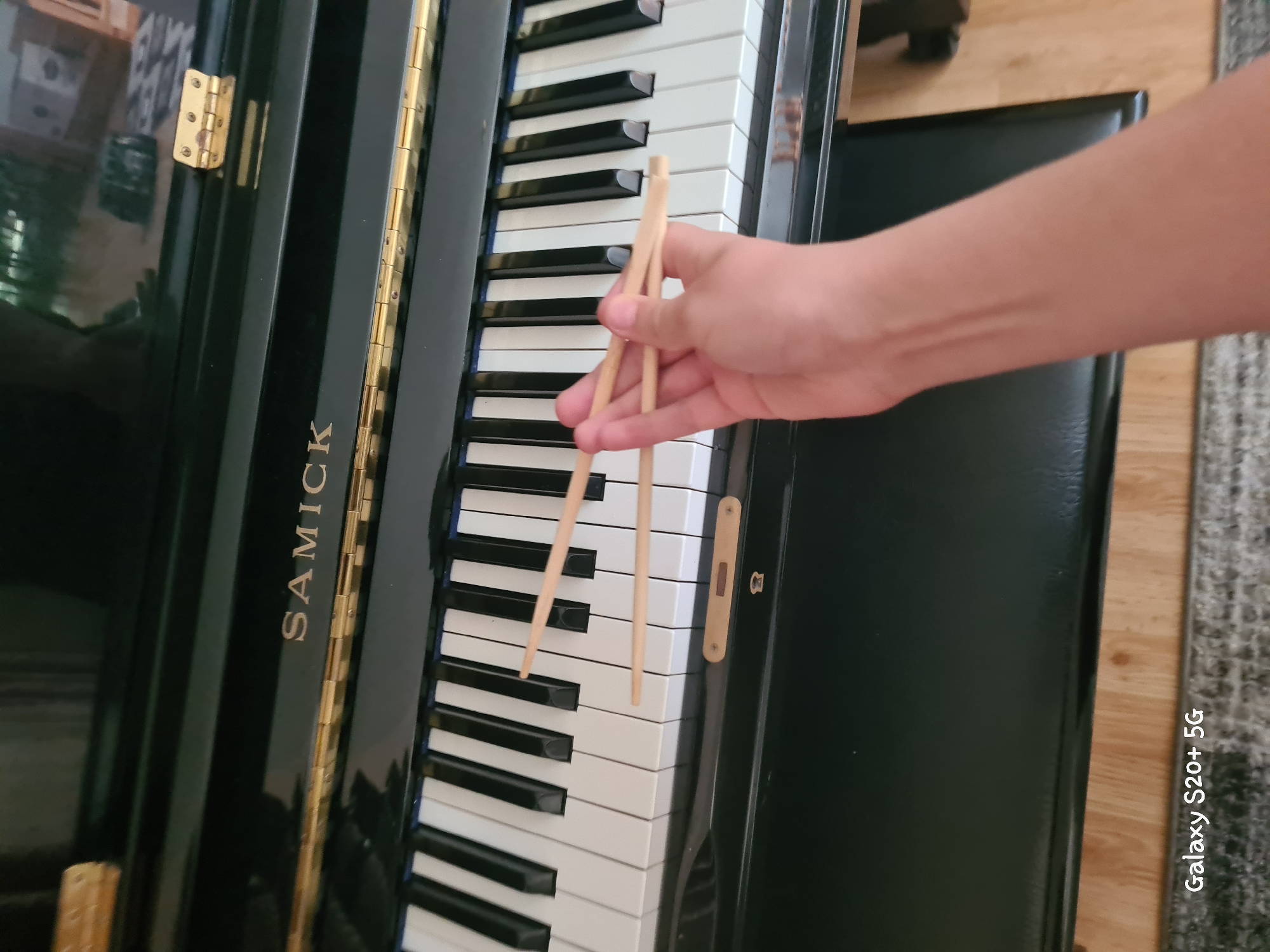An overhead shot shows a person's right arm and hand holding two wooden chopsticks over the black and white keys of a black upright piano. The "SAMICK" logo is visible on the piano's casing. The piano is situated in an indoor setting. A watermark reading "Galaxy S20+ 5G" is present in the bottom left corner of the image. There is no information to suggest the image was taken in Woodburn, United States of America.