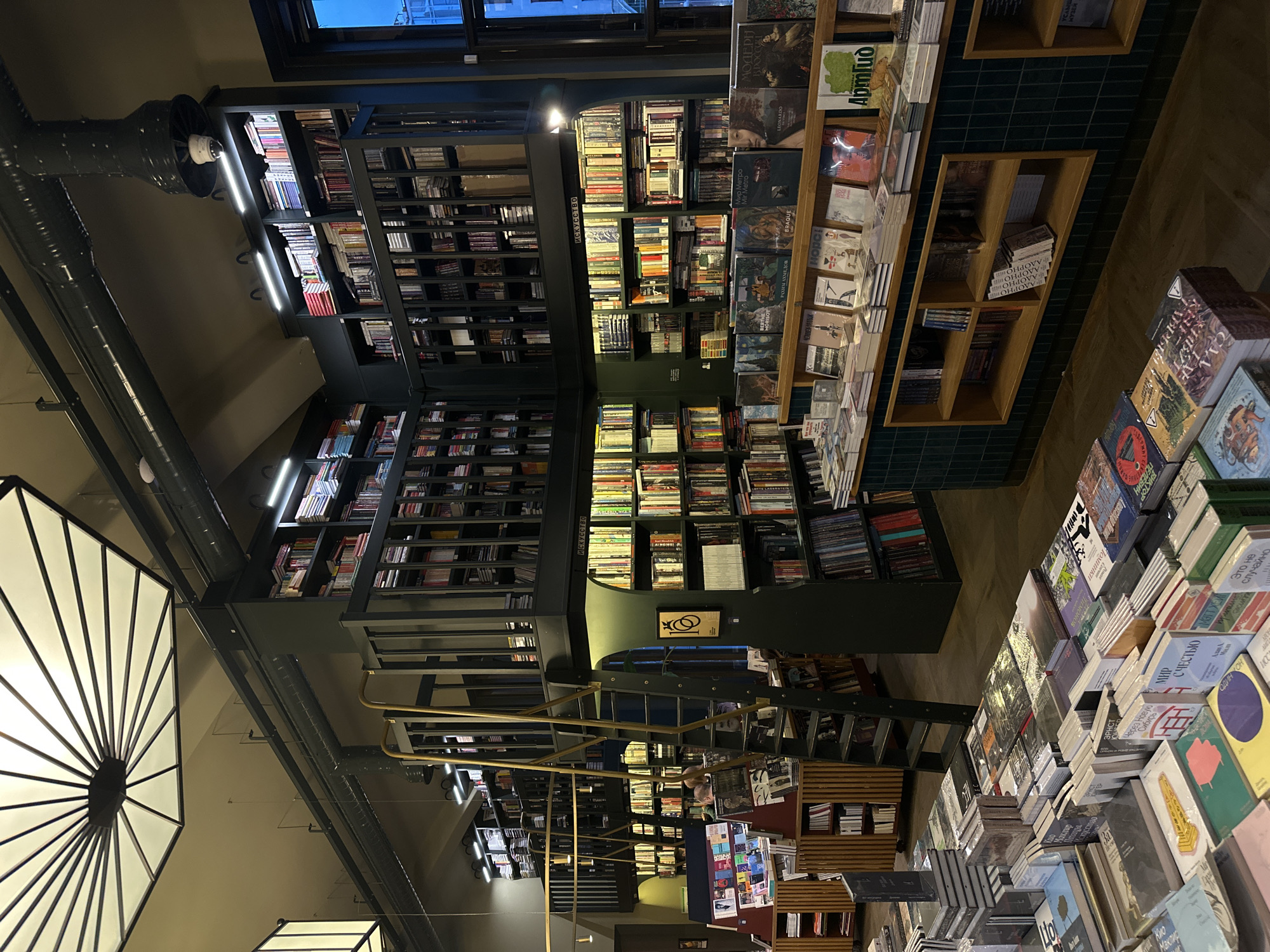 Here is a detailed description of the image:
The image shows the interior of a bookstore, likely in Unknown. The focus is on the bookshelves, which are packed with books. The shelves are of various heights and arranged in a multi-tiered structure. The books are stacked tightly, with many visible spines and covers in multiple languages.
The lighting in the store is a combination of natural light from windows and artificial light from overhead fixtures. The bookstore has a modern, loft-like design. Metal beams and ductwork are visible on the ceiling. A staircase with gold railings leads to upper levels of bookshelves.
The camera angle suggests the photograph was taken from a low vantage point, looking up at the books and the architecture of the store. The image is rotated counter-clockwise; the original image is rotated 90 degrees from its current orientation.