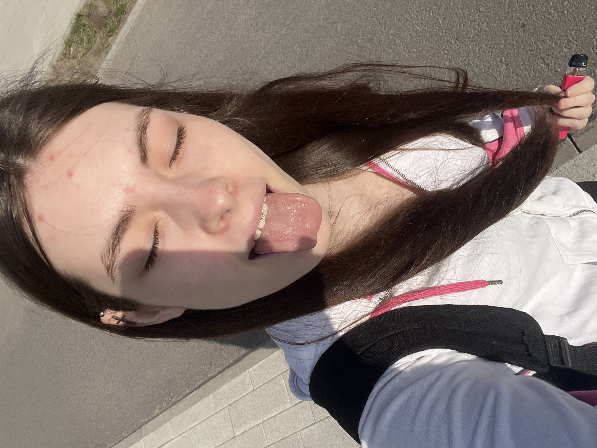 A young woman is pictured in a close-up shot. She has long, dark brown hair and is wearing a white jacket with pink accents. She is sticking her tongue out and her eyes are closed. In her hand, she is holding a pink vape device. The background appears to be a paved area. Given the architecture and lighting, it is likely that the photo was taken outdoors during the daytime. It is not possible to confirm that the photo was taken in the city of Saint Petersburg, Russia.