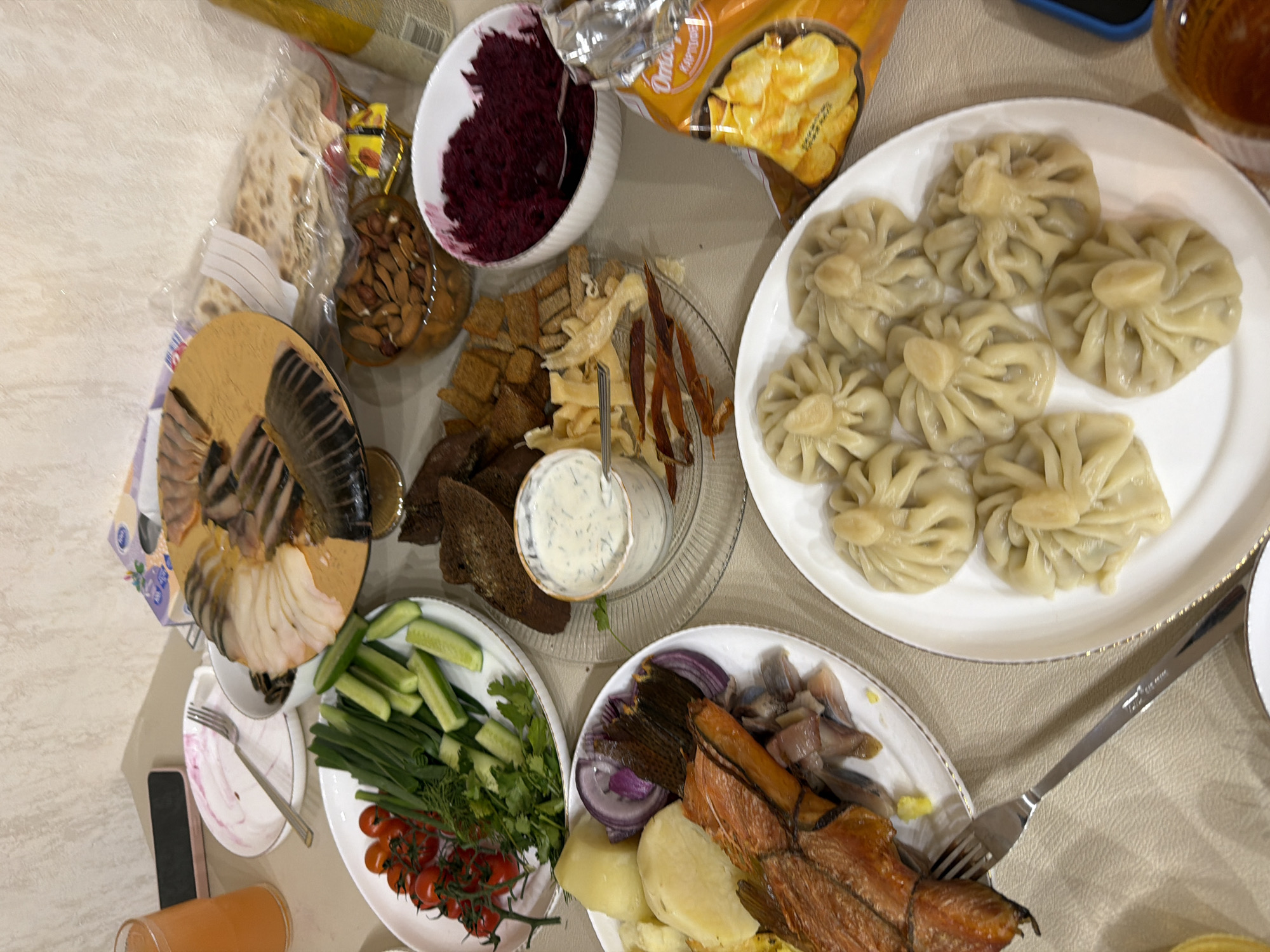 The image shows a table laden with food, likely a feast or celebration of some kind. In the top-right corner are khinkali, a type of Georgian dumpling, on a white plate. Beside it is a packet of potato chips. To the left of the khinkali is a white bowl filled with a dark-red beetroot salad. A glass plate with assorted types of bread, dried fish and small dish of white sauce is partially visible to the left of the beetroot salad.
A plate with sliced fish is also on the left side of the image. The lower left corner features a plate with sliced cucumbers, tomatoes, green onions, and dill, a white plate with sliced fish and a separate container with a pink liquid, likely some kind of juice. Another plate features herring, smoked fish, onions, and potatoes.
The scene takes place on a light-beige table. Based on the food choices (e.g., khinkali), it is likely a gathering celebrating Russian or Georgian culinary traditions. The exact location is not identifiable, but the reference to Chaikovsky, Russia, suggests that the image was taken there. No individuals are visible, so there are no interactions or actions directly discernible.