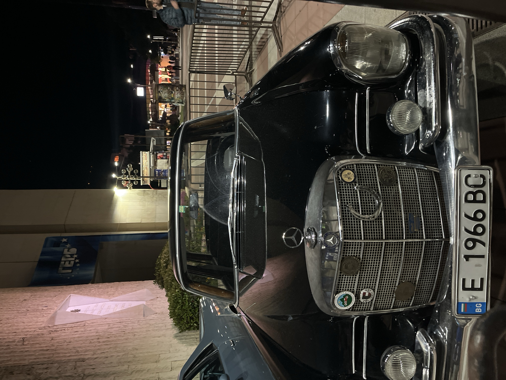 A front view of a black, vintage Mercedes-Benz car is captured at night, with a Bulgarian license plate reading "E 1966 BC" displayed prominently. The chrome grille of the car is adorned with several emblems and badges. The vehicle is parked in what appears to be a public area, indicated by the presence of streetlights and a metallic railing partially visible in the background. A person stands near the railing, in proximity to the vehicle. In the background, buildings and establishments are visible, with illuminated signs suggesting a commercial or urban environment. The setting seems to be Blagoevgrad, Bulgaria, based on the license plate’s country code and surrounding features.
