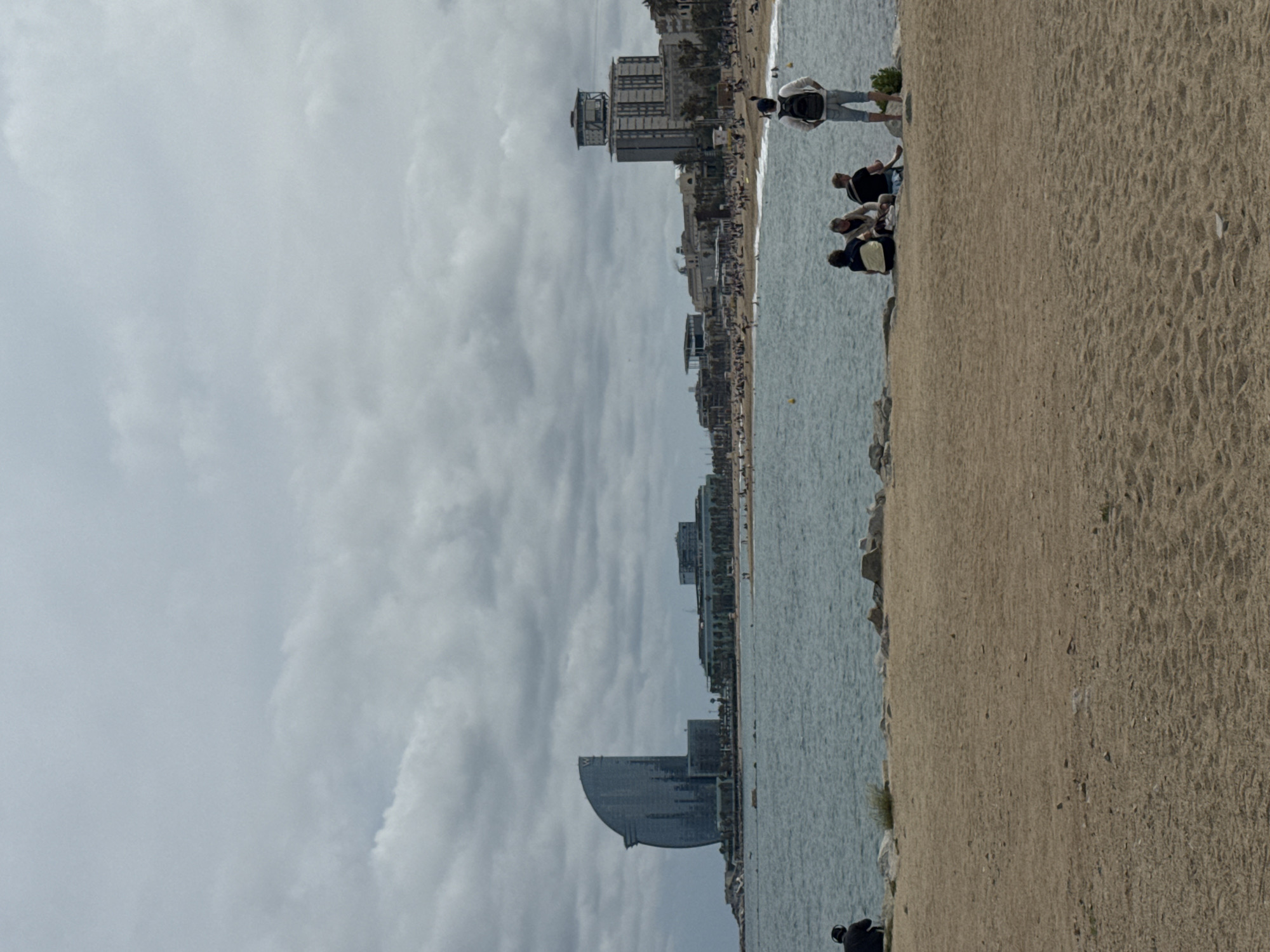 The image shows a view of the Barceloneta beach in Barcelona, Spain. The sky is cloudy. From the left, the W Barcelona hotel (the sail hotel) is visible. Various buildings line the coastline. The beach stretches along the water, with people visible walking, sitting, and standing near the water. A sandy area is in the lower right of the image.