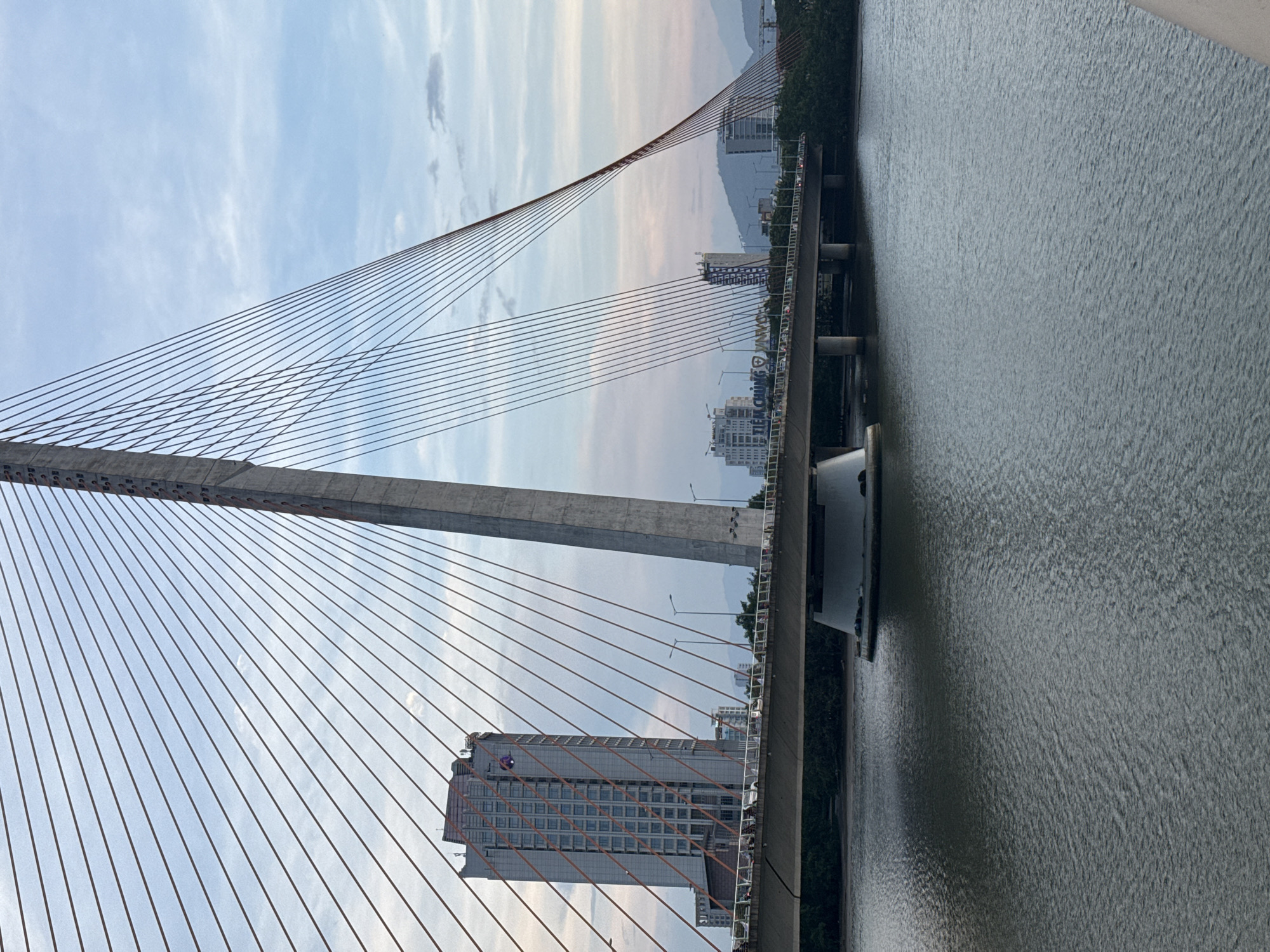 The image shows a view of the Dragon Bridge in Da Nang, Vietnam, from a distance. The bridge is a cable-stayed bridge spanning the Han River. The main pylon of the bridge is visible in the foreground, with its cables extending to the bridge deck. A tall building with a patterned facade is seen to the left of the pylon. In the background, other buildings, hills, and the sky are visible. The water of the Han River is in the foreground, with some ripples on its surface. The lighting suggests it is either early morning or late afternoon. There are no people or discernible actions captured in this static shot of the bridge and its surroundings.