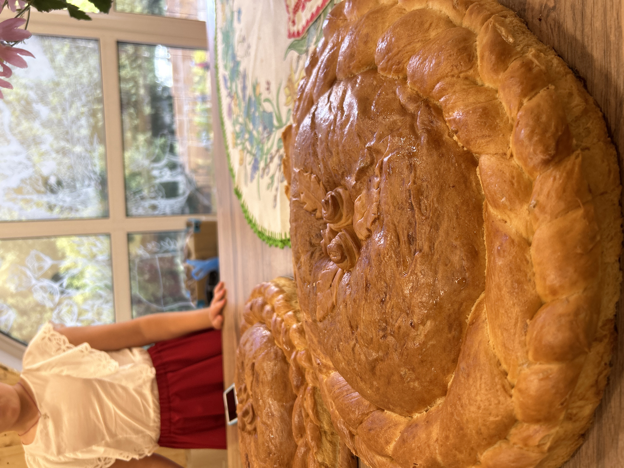 The image depicts a close-up view of two golden-brown, braided loaves of bread, likely traditional Karavai, placed on a wooden table. The foreground loaf is in sharp focus, showcasing intricate braided edges and decorative dough embellishments, including leaf-like patterns and rosettes. Behind it, another similar loaf is partially visible.
In the background, a young person, mostly out of focus, stands behind the table, wearing a white ruffled blouse and a red skirt. Their hands are resting on the table's edge. Beyond the table, a window is visible, offering a blurred view of greenery and trees, suggesting an outdoor or garden setting. A partially visible tablecloth with a floral pattern with green trim is on the table. The overall scene suggests a domestic setting, possibly a celebration or a special occasion.
Without additional information or context specific to Izhevsk, Russia, it is impossible to determine the precise location within the city. The image itself does not contain any identifiable landmarks or clues that would pinpoint its exact geographical placement in Izhevsk.
