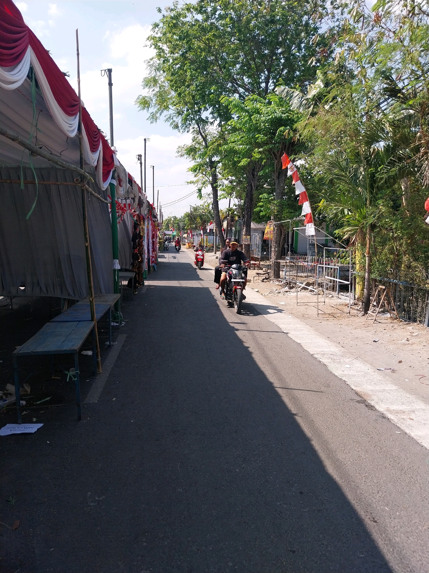 The image depicts a street scene in Campur, Indonesia, likely during a festive occasion given the red and white decorations. On the left side of the street, a large tent is set up with red and white draped fabric, suggesting a temporary structure for an event or market. Beneath the tent, there are several blue tables. The street itself is paved with asphalt, and there is a concrete curb on the right side. Two motorcycles are visible on the road. The first, further down the street, is red and is being ridden by a person wearing a helmet. Closer to the foreground, another motorcycle is also being ridden by two individuals. The rider wears a dark shirt and a yellow cap, while the passenger wears a dark shirt. Both are on a dark-colored motorcycle. Lush green trees line the right side of the street, and there are also some smaller plants and fences visible. Red and white flags are strung along the trees and a fence, further indicating a celebratory atmosphere. The sky is blue with scattered white clouds, and the sunlight casts shadows on the road, indicating it is daytime. Debris is scattered on the ground near the tables and along the roadside, with a piece of paper bearing the word "NGJUN" visible on the left.