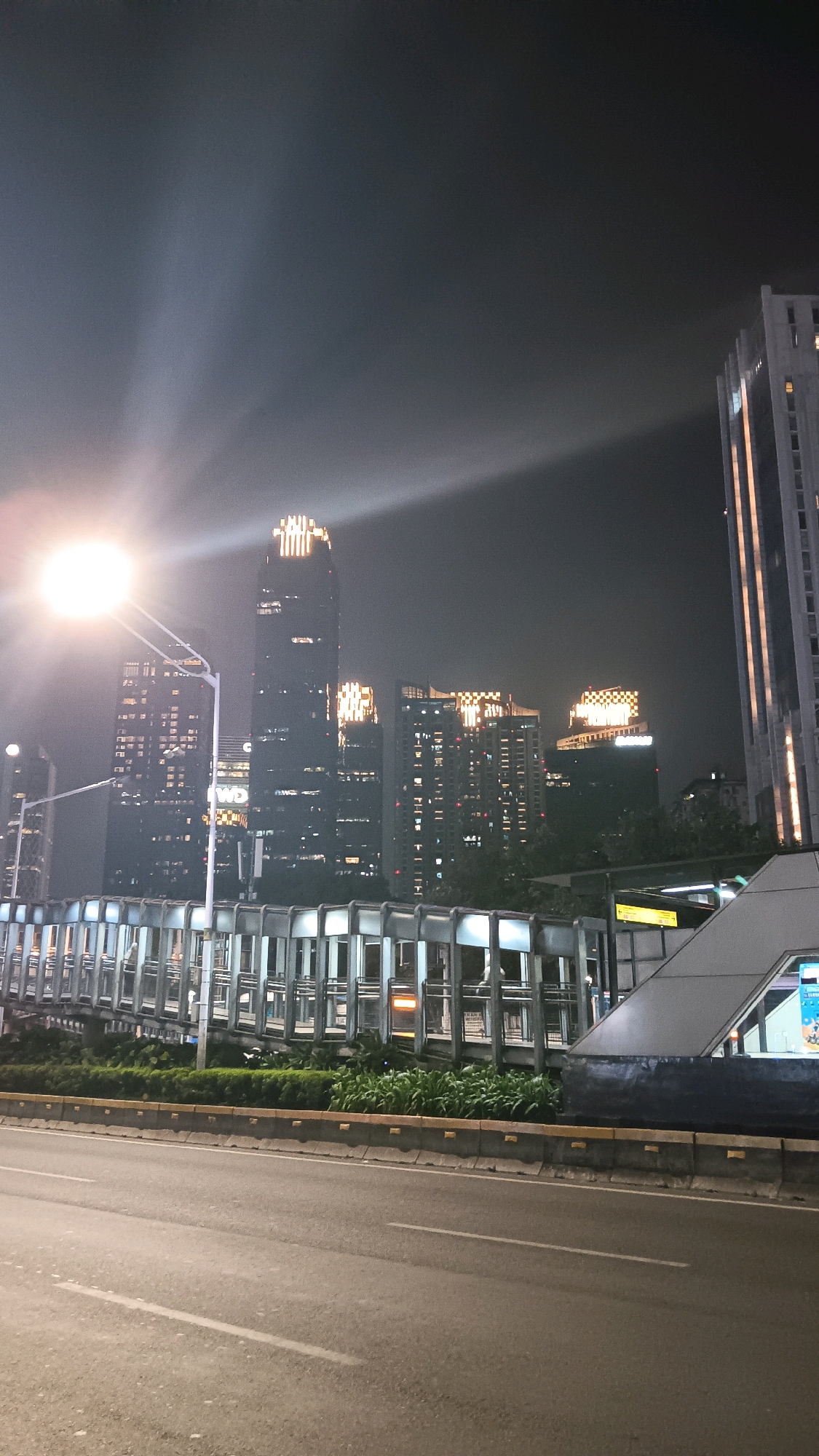 The image shows a nighttime view of the Jakarta skyline in South Jakarta, Indonesia, viewed from street level. The scene features a multi-lane road in the foreground, with concrete barriers and green foliage separating it from an elevated pedestrian walkway. The walkway is modern in design, with glass panels and a metal structure.
Behind the walkway, several tall buildings are illuminated against the dark night sky. The most prominent building on the left features a bright, glowing crown at its apex, with a large street lamp in the foreground casting a powerful, almost blinding light that streaks across the sky and over the buildings. Other skyscrapers in the background are also lit up, showcasing their windows and some illuminated signage or architectural features. One building in the mid-ground appears to have checkered patterns of light. The overall impression is of a vibrant, modern urban environment at night, with the city lights creating a dramatic contrast with the darkness. There are no discernible people in the image, and the focus is on the architecture and the cityscape.