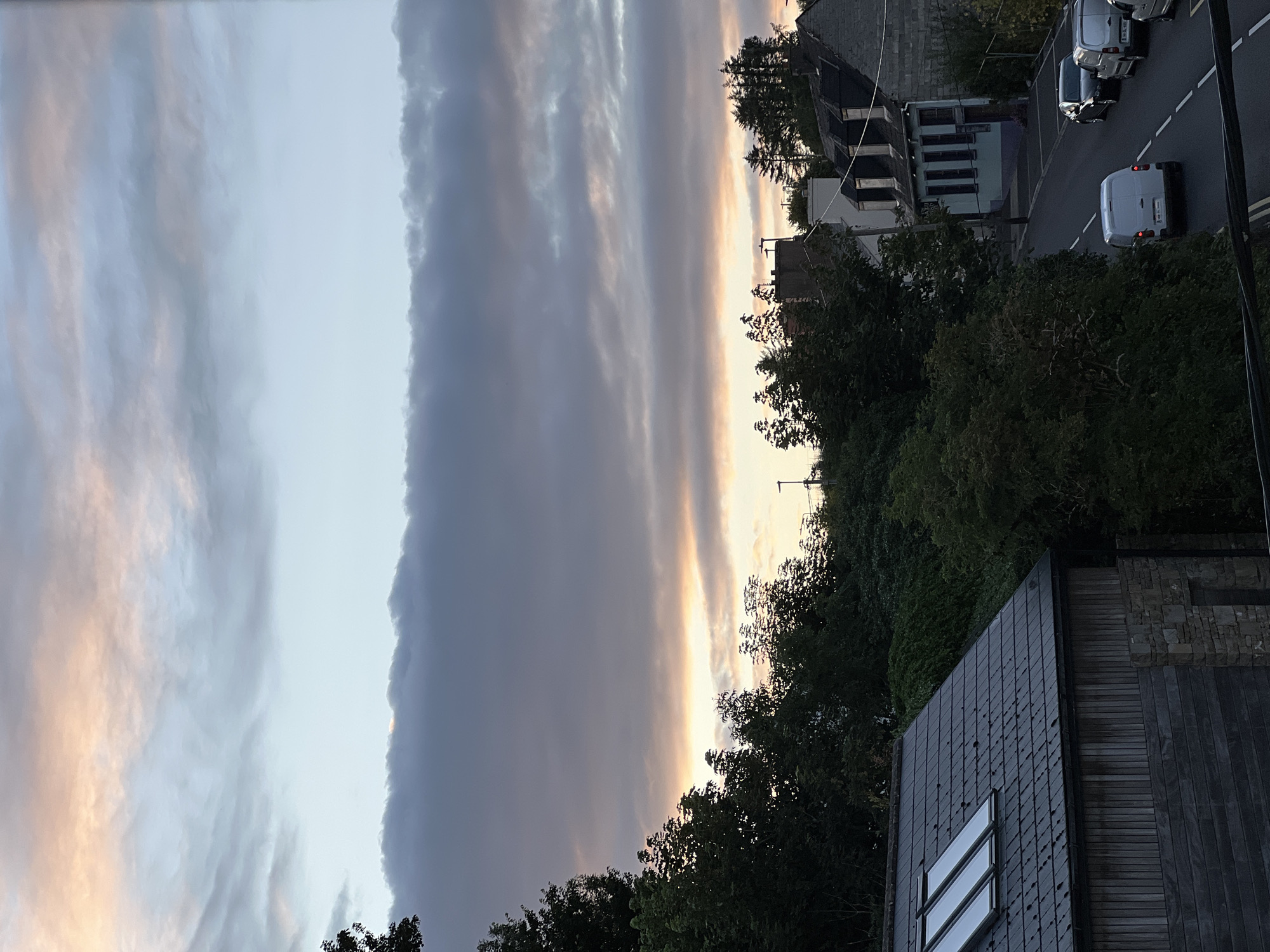 The image depicts a view of a street in Mountcharles, Ireland, at dusk. The sky is filled with dramatic clouds, tinged with pink and orange hues from the setting sun, contrasted with cooler blue and gray tones. In the foreground, the roof of a modern building with skylights is visible, constructed with dark tiles and wooden siding. To the right, a street curves uphill, lined with trees and several houses with pitched roofs. Various vehicles are parked and in motion on the street, including a white van, a dark car, and two other vans. Dense foliage and trees flank the left side of the street, obscuring much of the landscape behind them. The architecture suggests a residential or mixed-use area.
