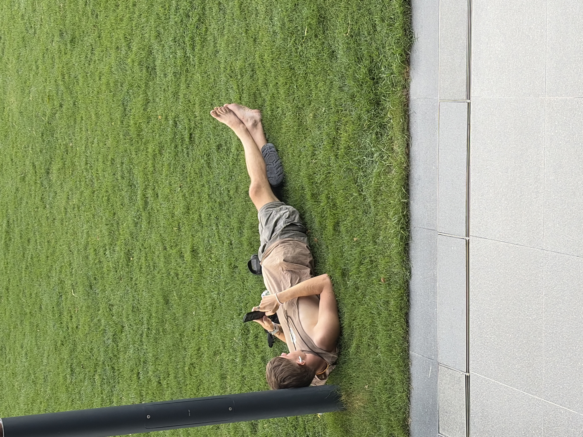 A man lies on green grass, with his bare feet pointing toward the top of the frame and his head toward the bottom. He wears khaki shorts and a tan shirt. He holds a cell phone in his hands and appears to be looking at it. White earbuds are in his ears. One shoe is on the ground by his legs. A dark gray pole is in the bottom left corner. Gray tiles are visible to the right. The photograph may have been taken in Bangkok, Thailand.
