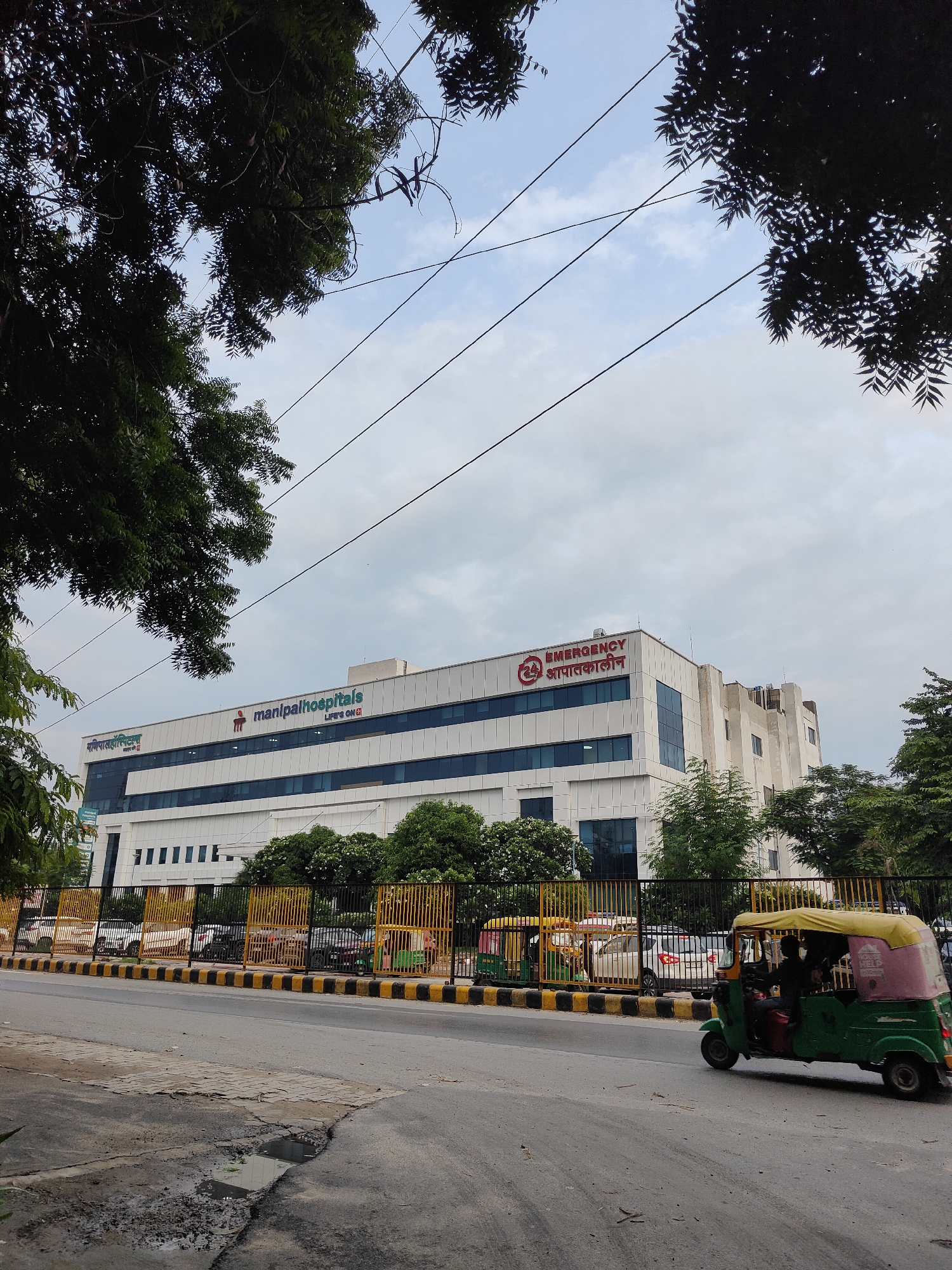 The image shows the Manipal Hospitals in Gurgaon, India, on a cloudy day. The hospital building is a modern, multi-story structure with white cladding and blue-tinted windows. The "manipalhospitals" logo is visible on the building, along with the text "LIFE'S ON." A section of the building also displays "EMERGENCY" in red letters, with the Hindi translation "आपातकालीन."
In the foreground, a street with a yellow and black striped border runs along the front of the hospital. Several auto-rickshaws are on the road, with one in the right foreground being green and pink. A black metal fence with yellow vertical bars separates the street from the hospital property, behind which cars and more auto-rickshaws are parked. Trees, some with dark green foliage, are visible on the left and right sides of the image, as well as in front of the hospital. The sky is overcast with some faint outlines of clouds. The road surface shows some signs of wear and tear, including patches of broken asphalt and puddles of water.