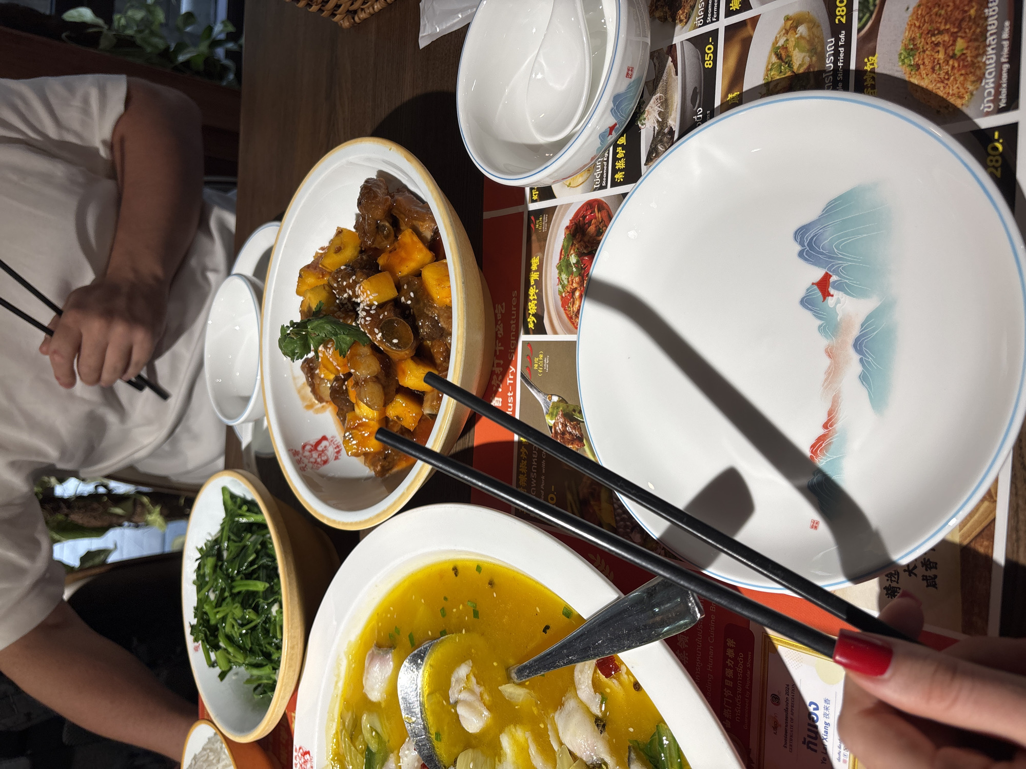 Two individuals are dining at an indoor restaurant, featuring a wooden table surface and warm artificial lighting, suggesting an evening meal. The setting appears to be a Chinese or Hunan cuisine establishment, confirmed by text on the laminated menus/placemats spread across the table.
On the left, a person wearing a white shirt is visible, holding black chopsticks. On the right, another person's hand with red nail polish is also holding black chopsticks, positioned over an empty white plate.
Several dishes are laid out for sharing. These include a large white bowl containing sweet and sour meat (possibly pork or chicken) with mango or pineapple chunks, garnished with sesame seeds and cilantro. Another bowl holds green leafy vegetables, likely stir-fried. A prominent oval dish features fish fillets immersed in a vibrant yellow, soupy sauce with green onions and red specks, accompanied by a large serving spoon. Empty white ceramic bowls with a blue rim and white ceramic plates adorned with an artistic blue and red mountain/landscape design are also present.
The menus display a variety of dishes with corresponding prices, written in both Thai script and Chinese characters, with some English translations. Notable visible text includes "Leading Hunan Cuisine Br", "Guaranteed by Popular Shows", "CERTIFICATE OF APPRECIATION", and the restaurant name "Yo Lai Xiang 夜来香". Specific dishes mentioned are "Yelaixiang Fried Rice" (ข้าวผัดเย้หลายเซีย), "Steamed Egg" (ไข่ตุ๋นกุ้ง), "Steamed Seabass" (清蒸鲈鱼), and "Must-Try Signatures". The prices shown are 850.- and 280.- Thai Baht. The presence of Thai script, coupled with the provided context, indicates the location is Bangkok, Thailand.
