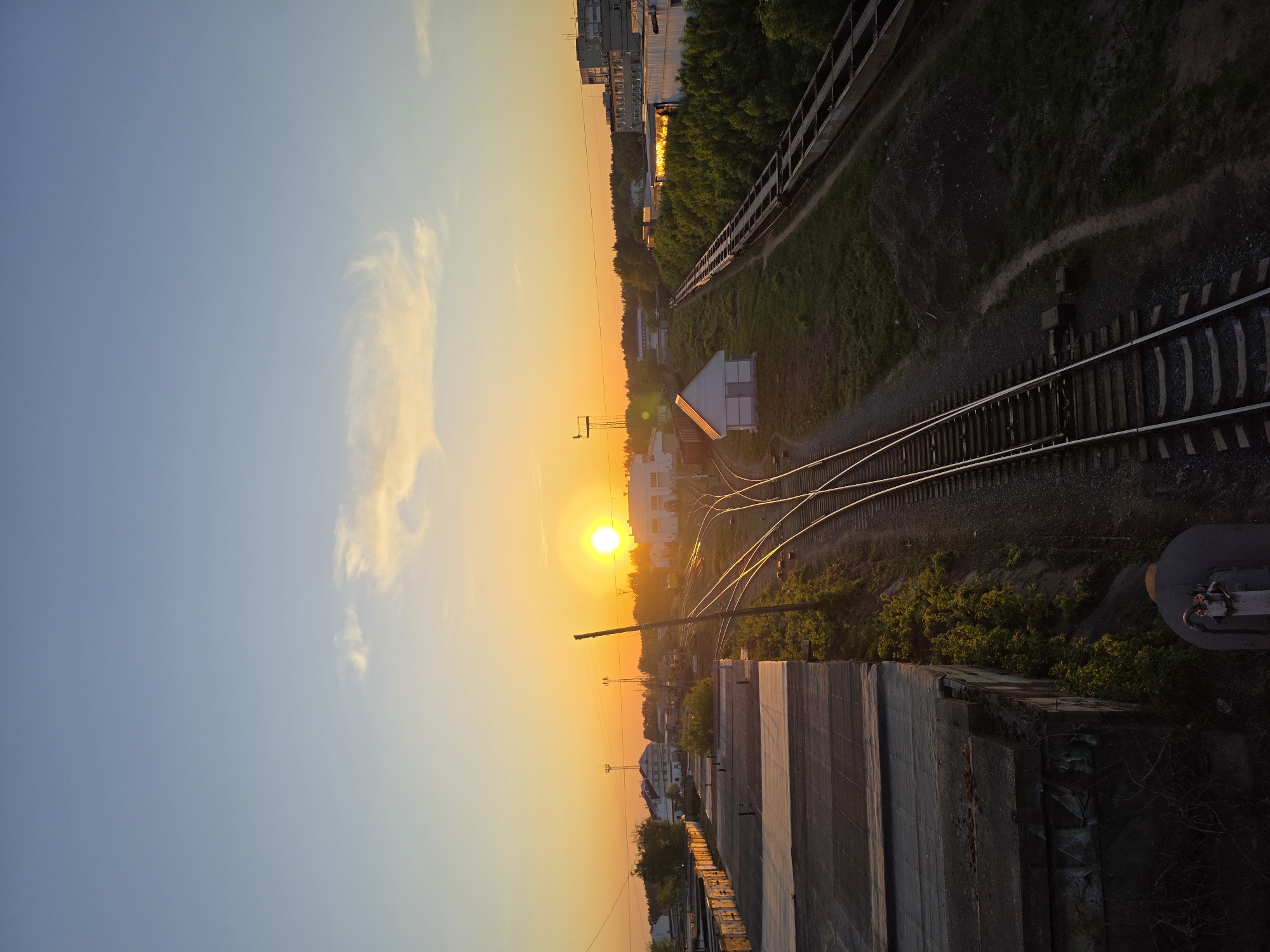 A vertical, eye-level view captures a scene near train tracks in Chaikovsky, Russia, illuminated by a bright setting sun. The sky transitions from a light blue in the upper left to a warm orange hue near the horizon.
Dominating the right side of the image are train tracks, with multiple lines converging in a switchyard. Vegetation and trees line the tracks. In the center of the image, the bright sun creates a lens flare effect, obscuring details in the immediate vicinity. Light reflects off the top of a white building. Utility poles and lines can be seen silhouetted against the sun.
The lower portion of the image features a concrete structure, possibly a bridge or retaining wall, its surface worn and textured. In the upper-right, buildings appear in the distance. The general environment appears industrial or urban, with the focus on the railway infrastructure and the sun as the dominant light source.