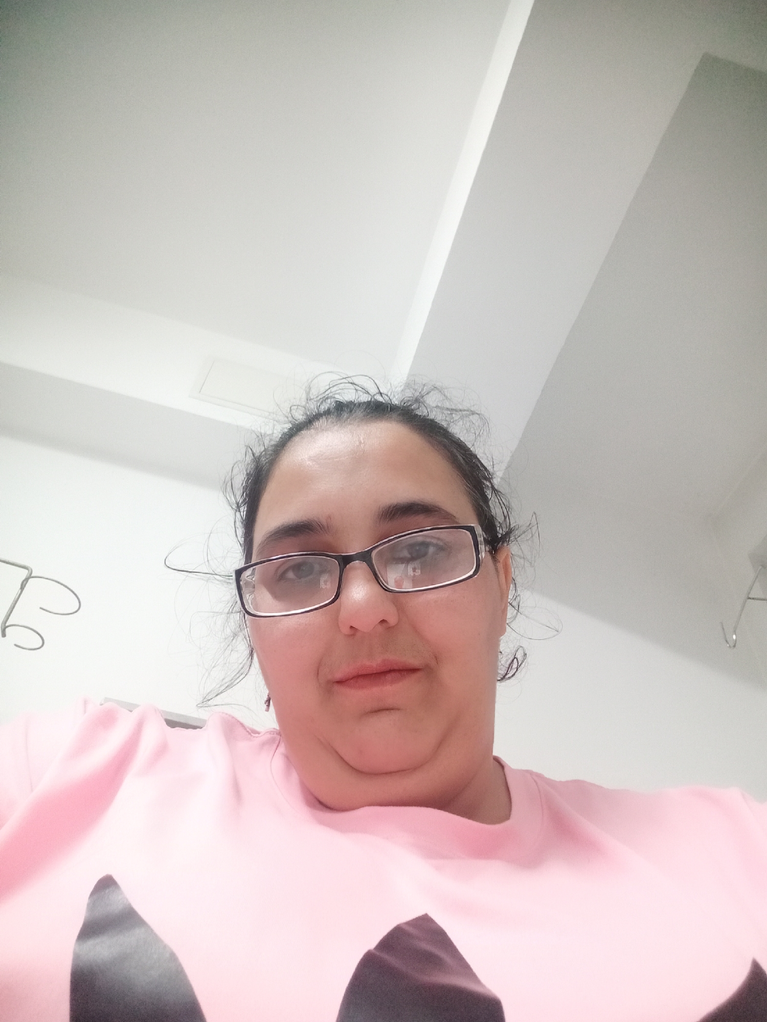 The image shows a close-up, frontal selfie of a woman with dark hair and glasses, wearing a pink sweatshirt. She is looking directly at the camera. The background is a white ceiling with visible architectural details. The image does not contain any identifiable elements related to the city of Pleven, Bulgaria.