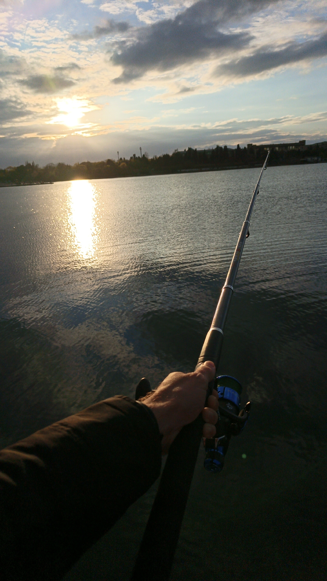 A first-person perspective shot shows a person holding a fishing rod over a body of water, possibly the Black Sea in Sevastopol, Ukraine. The sun is setting or rising on the horizon, its light reflecting on the water's surface. The sky is filled with clouds. Across the water, a dark landmass is visible, possibly a shoreline with buildings and vegetation. The fishing rod is the prominent object, with the person's hand and arm holding the rod close to the fishing reel.