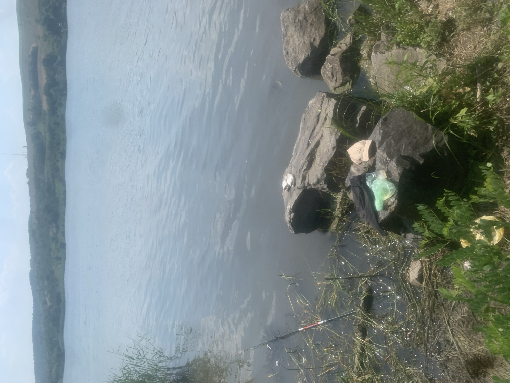 A fishing rod rests on the bank of a calm, murky river in Kosy, Ukraine. The water is still, with a few ripples disturbing its surface. On the rocky bank, a black cloth and a light green plastic bag lie near some large stones. Lush green vegetation grows around the rocks, with dry grass and some fallen leaves also visible. In the distance, a gently sloping, tree-covered hill forms the opposite bank of the river. The sky above appears hazy or overcast, suggesting a mild day. The scene conveys a sense of quiet solitude, with the focus on the peaceful activity of fishing. No people are visible, and there is no discernible text in the image.