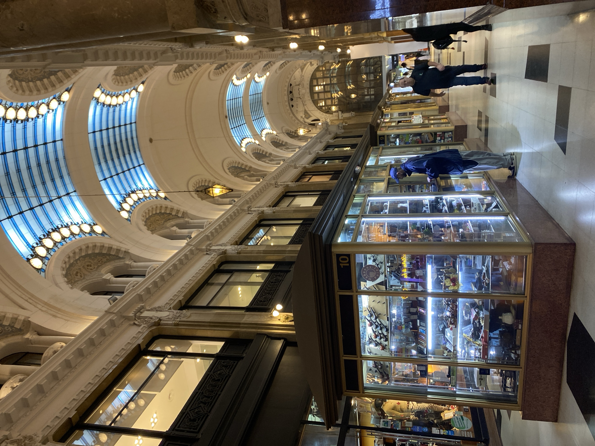 The image depicts a view inside a historical shopping arcade in Buenos Aires, Argentina. The architecture features a barrel-vaulted ceiling with a blue and white striped glass skylight illuminated by rows of light bulbs. The walls are adorned with decorative moldings and pilasters.
Along the arcade, storefronts are visible, with display cases filled with various items. Two men are present in the scene: one is peering into a display case, and another man is walking in the opposite direction. The flooring consists of square tiles in a black and white pattern. The atmosphere is well-lit, highlighting the ornate details of the arcade.