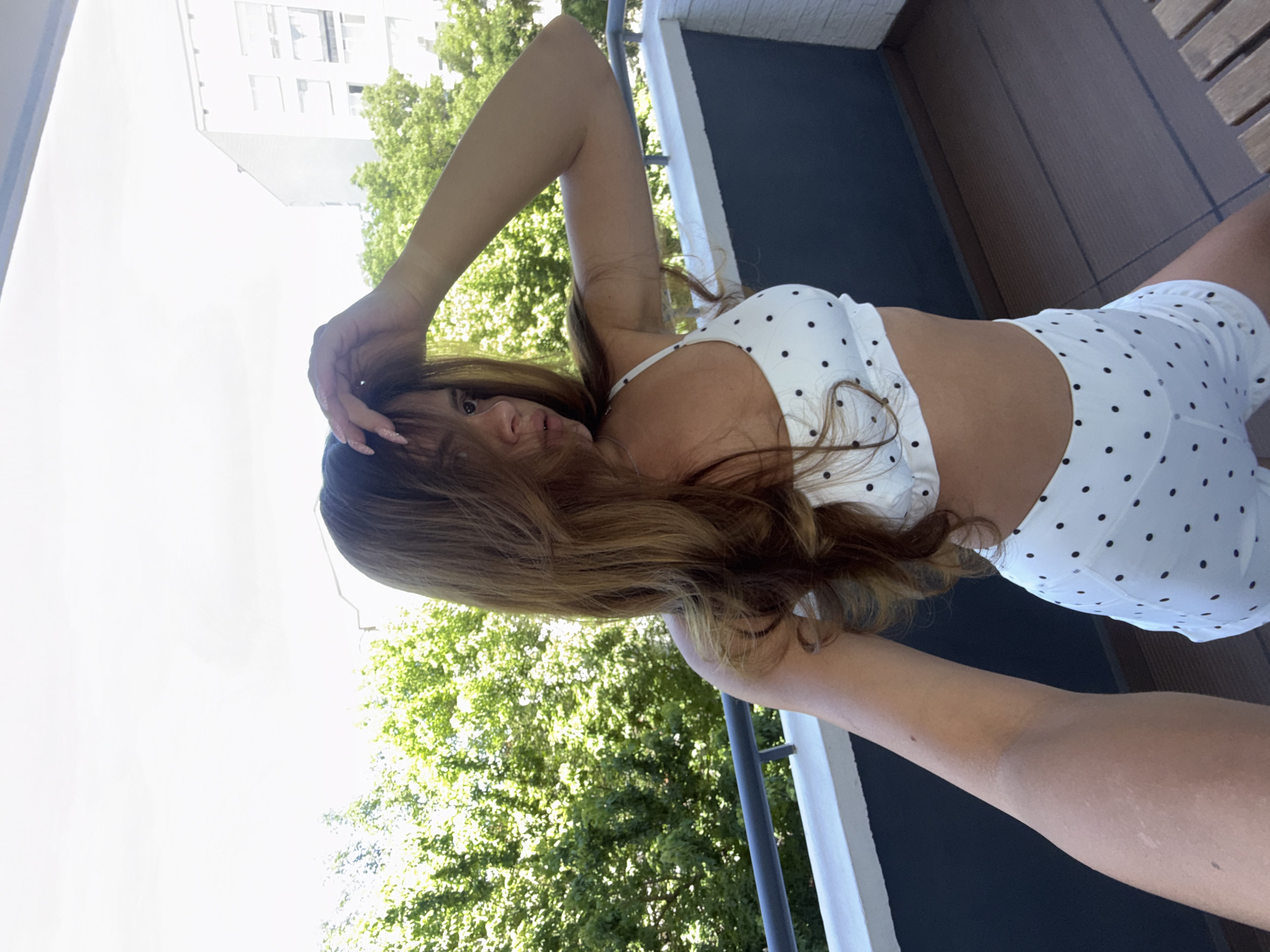 The image shows a young woman with long, light brown hair standing on a balcony in Plovdiv, Bulgaria. She is wearing a white polka dot bikini top and matching shorts. Her left arm is raised, and she is touching her hair. The balcony has a railing and a wooden floor. In the background, there are green trees and a building. The sky is overcast.