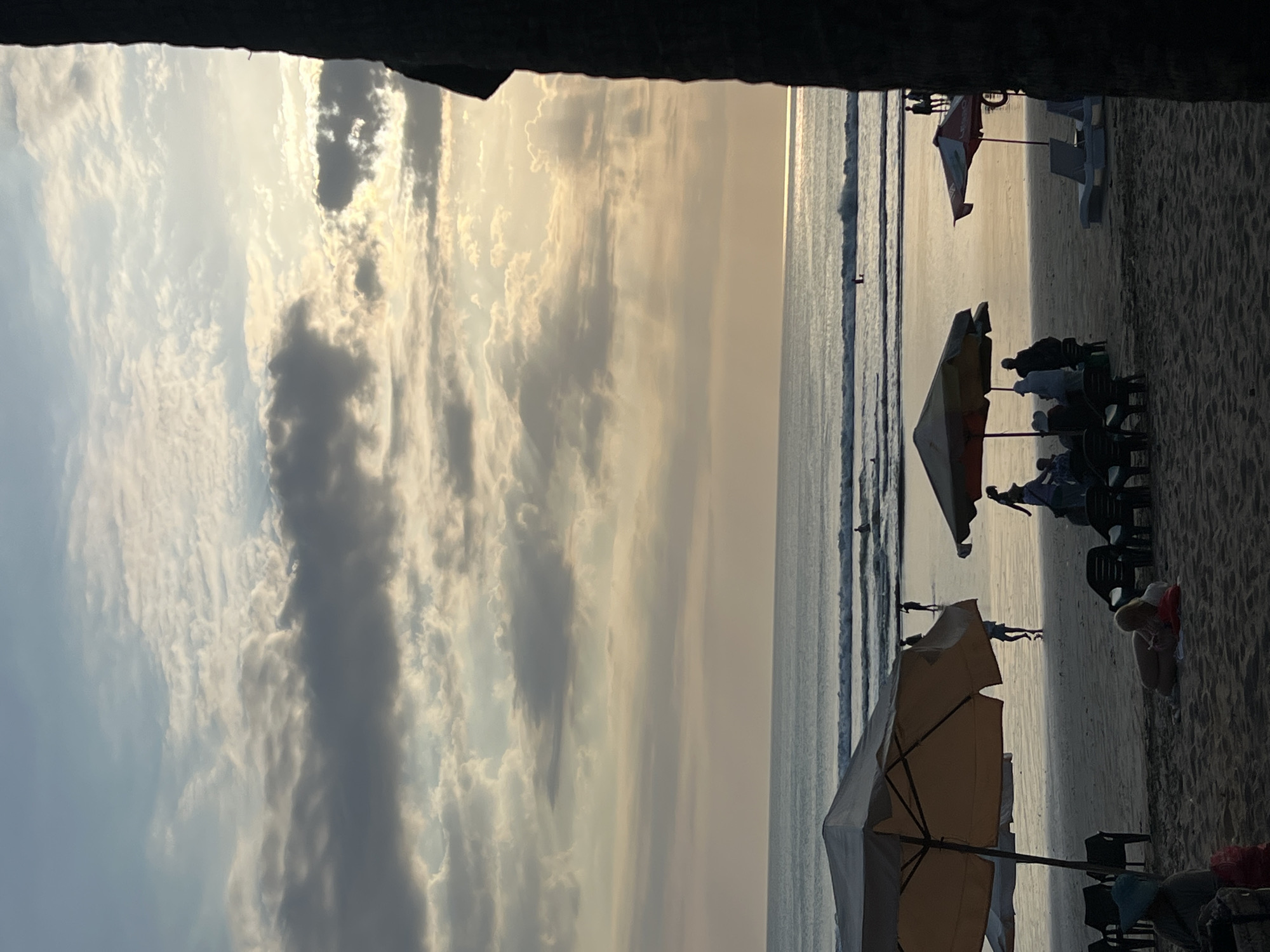 This is a medium shot of Kuta Beach in Bali, Indonesia at sunset. The sky is filled with dramatic clouds, with rays of sunlight breaking through. The ocean is calm with gentle waves, and a few surfers are visible in the distance. The beach is sandy, with several beach umbrellas and lounge chairs set up. People are seen walking along the shore and relaxing under the umbrellas. The overall atmosphere is peaceful and serene, with the golden hues of the sunset casting a warm glow over the scene.
