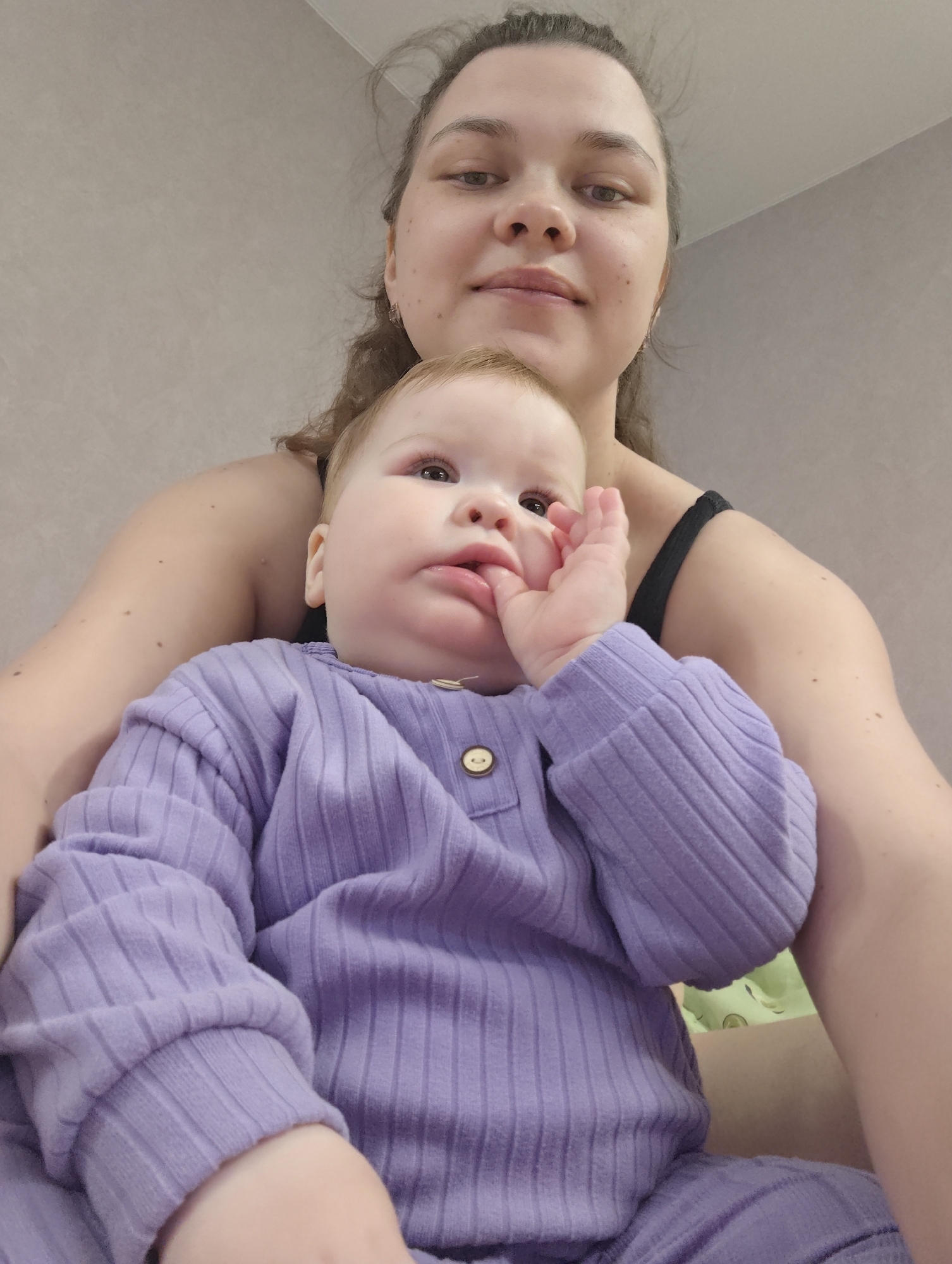 A medium shot captures a mother and her baby. The mother, with her hair pulled back, faces the camera with a soft expression. She's wearing a dark-colored tank top, and her arms gently encircle the baby. The baby, dressed in a ribbed, button-down, purple outfit, sits comfortably in the mother's lap. The baby's hand is near its mouth, possibly sucking on its fingers. The background is a plain, light-colored wall, suggesting an indoor setting. The location is the city of Chaikovsky, Russia.