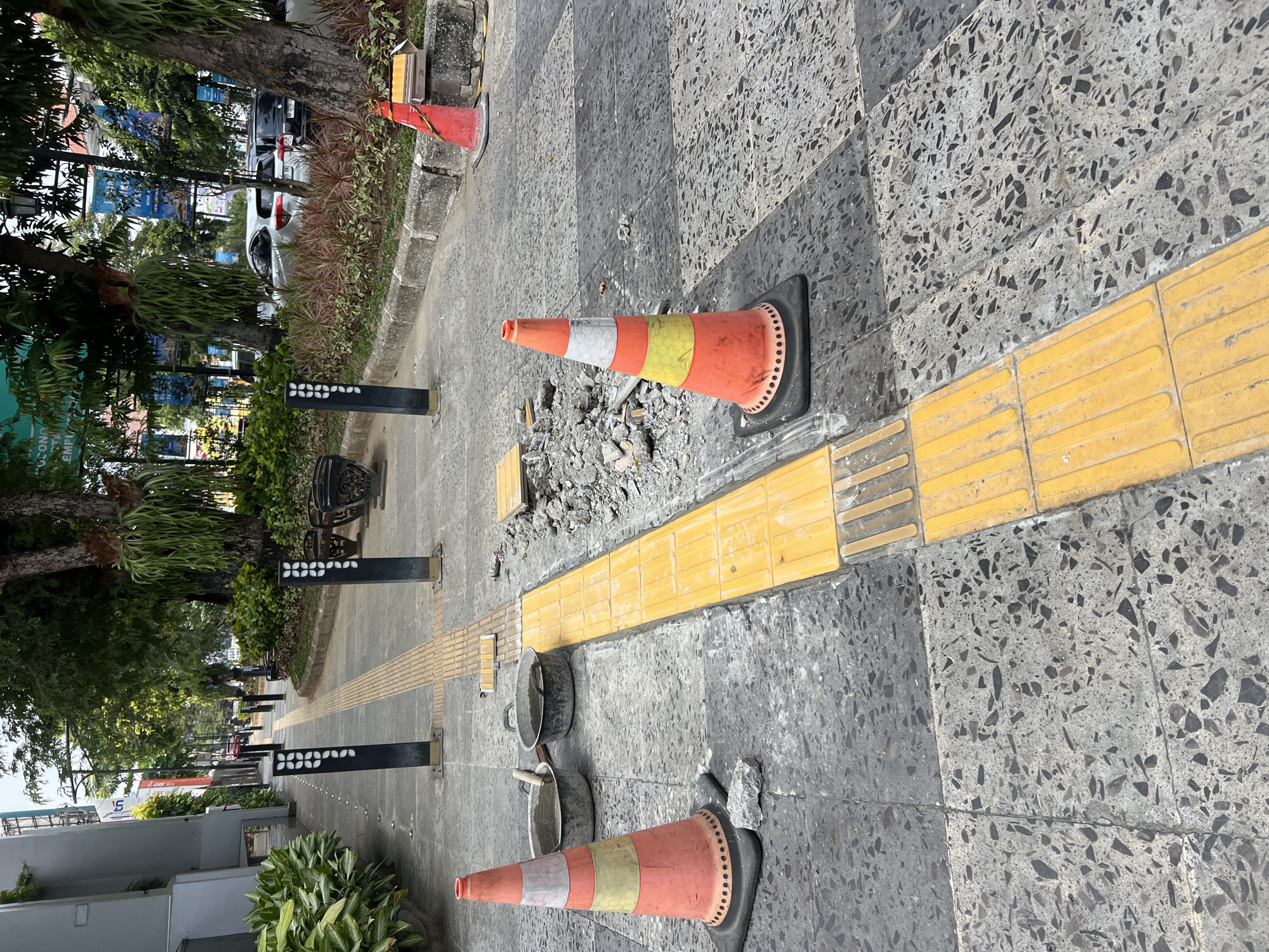 The image depicts a section of a city sidewalk, specifically in Yogyakarta, Indonesia, undergoing repair. The sidewalk is paved with gray speckled tiles, and a raised yellow tactile paving strip, designed for visually impaired pedestrians, runs along its length. Construction debris, including broken concrete and a yellow paving tile, is scattered on the ground. Two orange traffic cones are placed to demarcate the work area. In the foreground, two gray buckets, one with a wooden handle, contain a grey mixture, likely cement or grout, indicating ongoing construction. In the background, trees and lush green foliage line the sidewalk, and beyond them, parked cars are visible on a street. Several black bollards with white decorative elements are also present along the sidewalk. The overall scene suggests a typical urban environment where infrastructure maintenance is in progress.