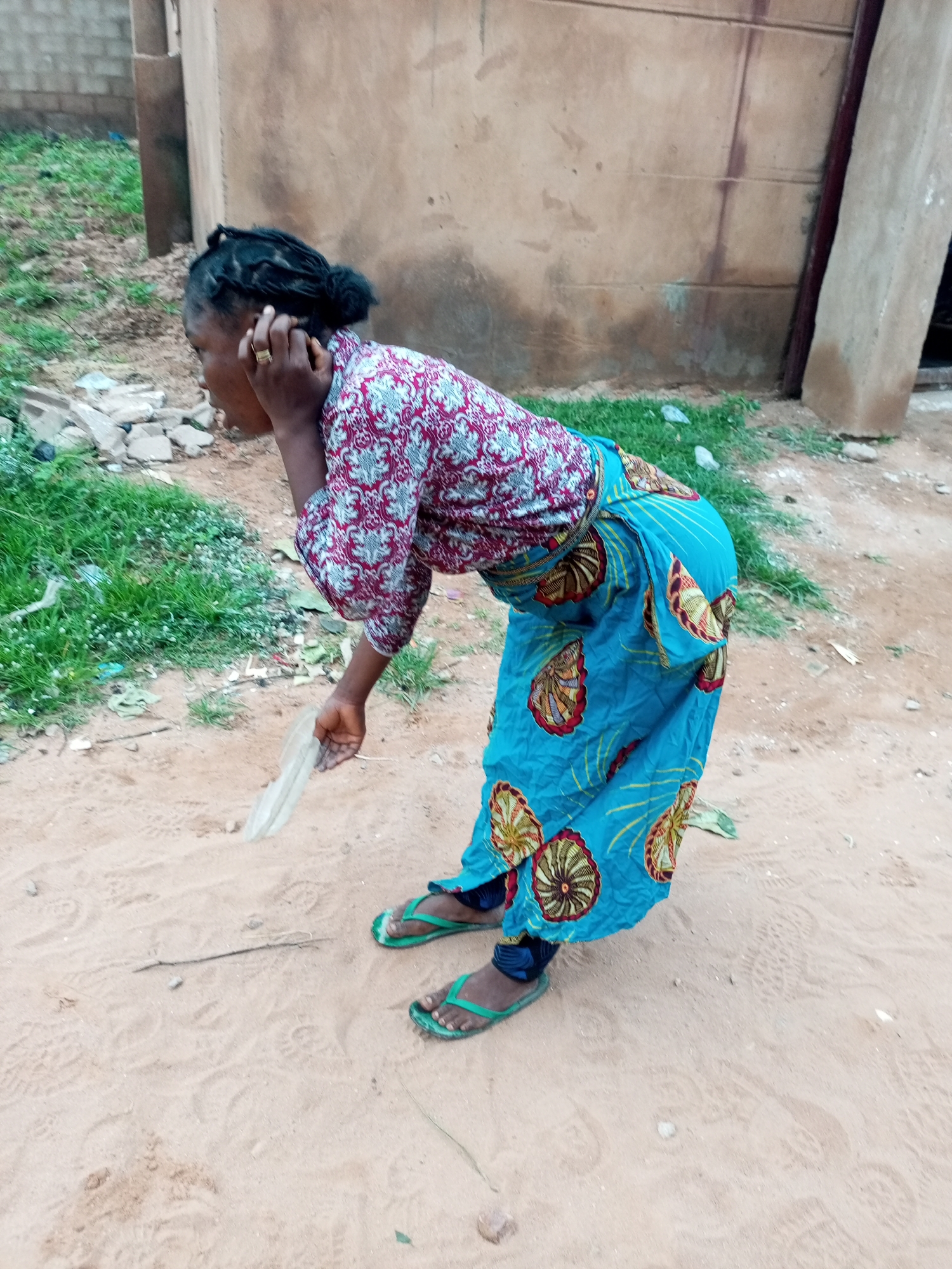 A woman with dark skin and braided hair is bent over in an outdoor setting. She is wearing a red and white patterned long-sleeved shirt and a blue skirt with a yellow and red circular pattern. Her right hand is touching her ear, and she is holding a thin, light-colored object, possibly a piece of paper or plastic, in her left hand. She is wearing green flip-flops. The ground is sandy and has some grass and debris on the left side of the image. In the background, there is a textured wall. The scene appears to be in a rural or semi-urban area in Tempure, Nigeria.