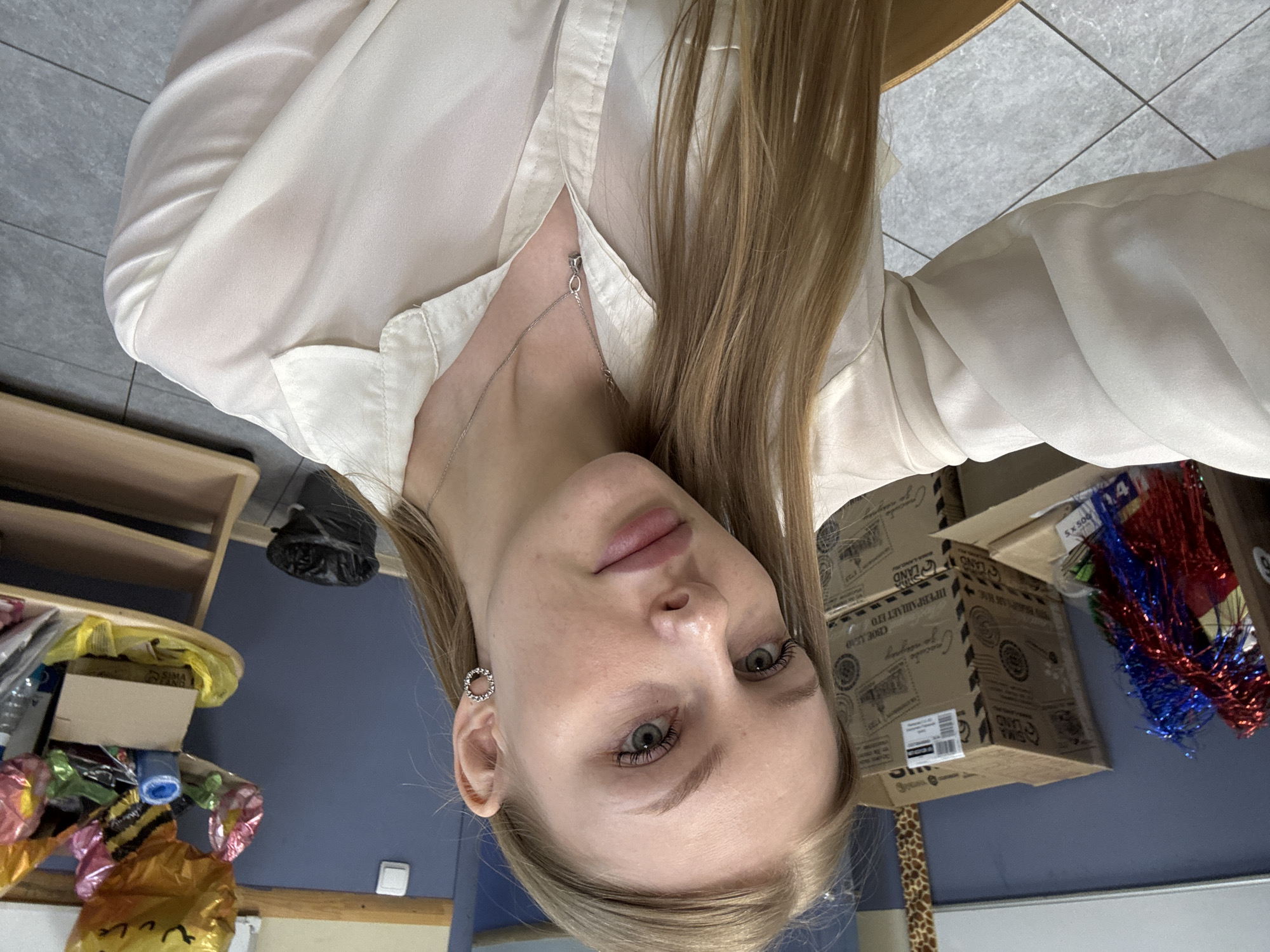 A fair-skinned, blond woman is the focal point of the image, viewed from a slightly rotated close-up perspective. She has fair skin, green eyes, and light eyebrows and eyelashes. She wears a silver chain necklace and circular silver earrings. Her hair is long, straight, and parted off-center. She is wearing a light-colored blouse, partially unbuttoned.
The background contains several objects:
* Boxes with Russian text, possibly packaging materials, are stacked. The text includes the word "LAND".
* Red, white, and blue tinsel or decorations are visible, suggesting a festive or party-related event.
* A shelf with various items is present, including balloons and boxes.
The location cannot be definitively confirmed as Perm, Russia, based on the visual information alone, although the Russian text on the boxes suggests a location in Russia.