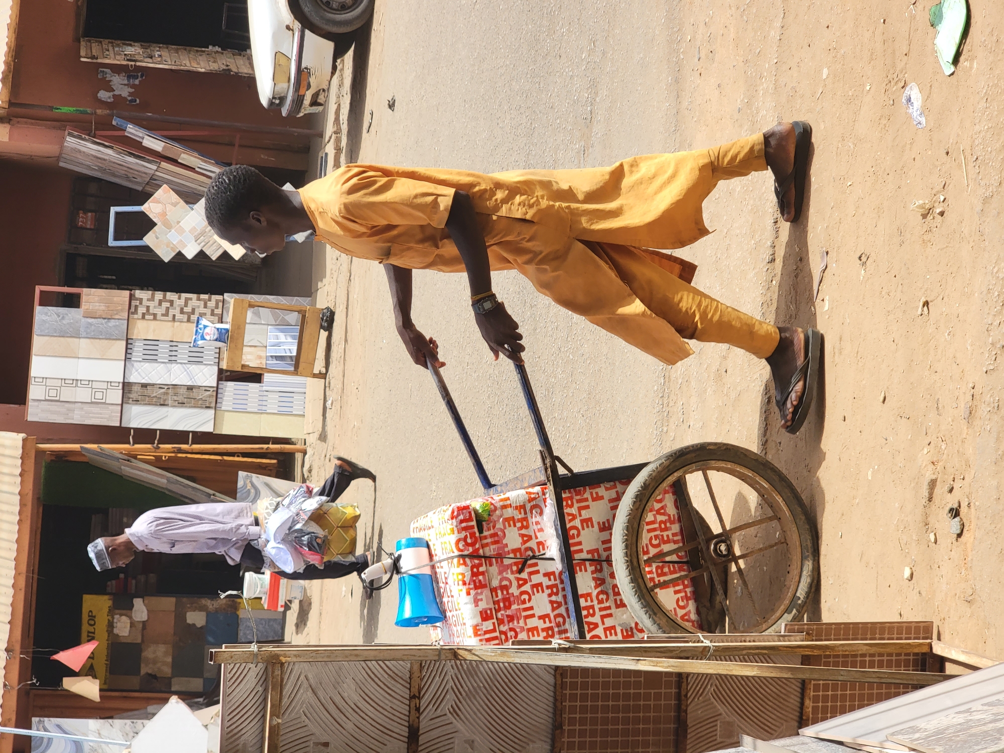 In a street scene likely located in Maiburiji, Nigeria, a dark-skinned man in a yellow outfit pushes a two-wheeled cart laden with packages marked "FRAGILE". He wears sandals and a wristwatch. In the background are shops displaying tile samples, suggesting a commercial district. The street is dirt, and sunlight casts harsh shadows. Another man carrying bags, likely a customer, is seen walking in the background near one of the shops. A white car is also partially visible.