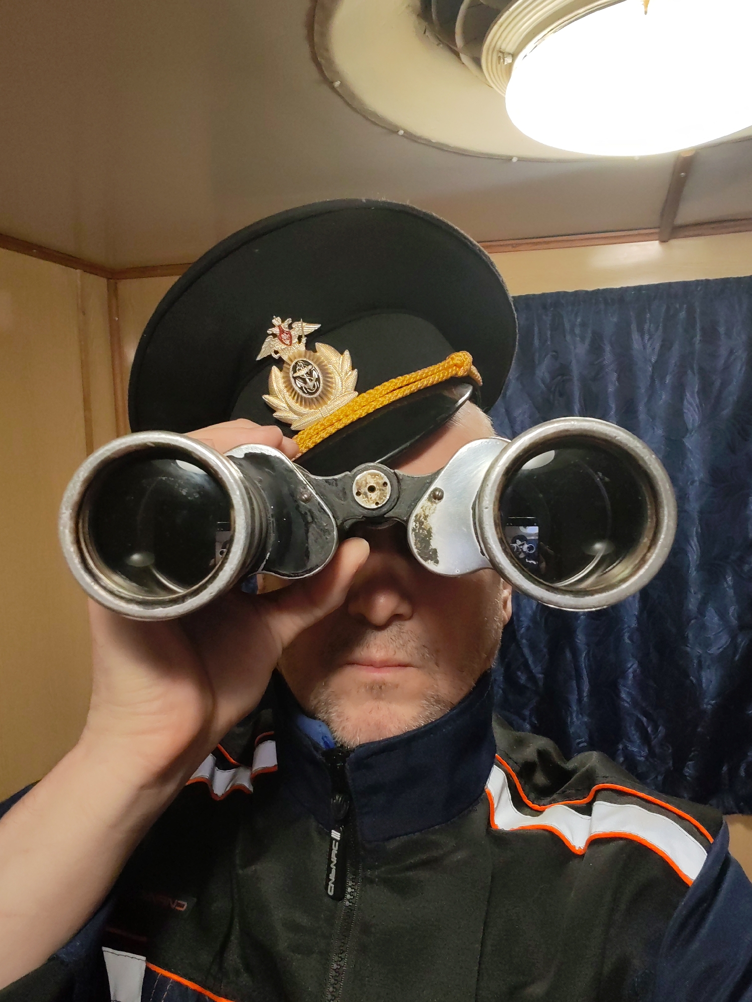 A man in Rybinsk, Russia, is captured in a waist-up portrait, holding a pair of metallic, vintage binoculars to his eyes, as if looking through them. He wears a black uniform peaked cap featuring a gold cord and an elaborate emblem with a double-headed eagle, anchors, and a circular motif, indicative of Russian maritime or military insignia. The man has light facial hair and a focused, neutral expression.
He is dressed in a dark blue or black jacket with prominent orange and white stripes along the shoulders and arms, resembling reflective detailing. The zipper pull on his jacket bears the text "CNNAC".
The setting is an indoor space, likely a ship's cabin or similar functional area, characterized by a curved, off-white ceiling and light brown wooden wall paneling. A circular, illuminated light fixture is visible on the ceiling. Dark blue, textured curtains are partially visible in the background to the right. The overall impression is one of observation or duty within a maritime or industrial context.