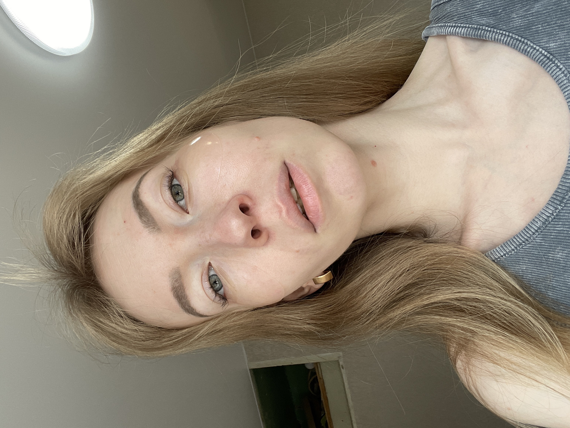 A young woman with long blonde hair and fair skin is posing for a selfie. She is wearing a gray ribbed tank top and a gold hoop earring. Her eyes are a gray-green color, and she has light brown eyebrows. Her lips are a pale pink. There are a few blemishes on her face. The background is a neutral gray color, with a light fixture visible in the upper left corner. A partially visible doorway is present in the lower part of the image. The photo appears to have been taken indoors, with a sideways, tilted perspective. There is no specific evidence in the image to suggest a location in the city of Perm, Russia.