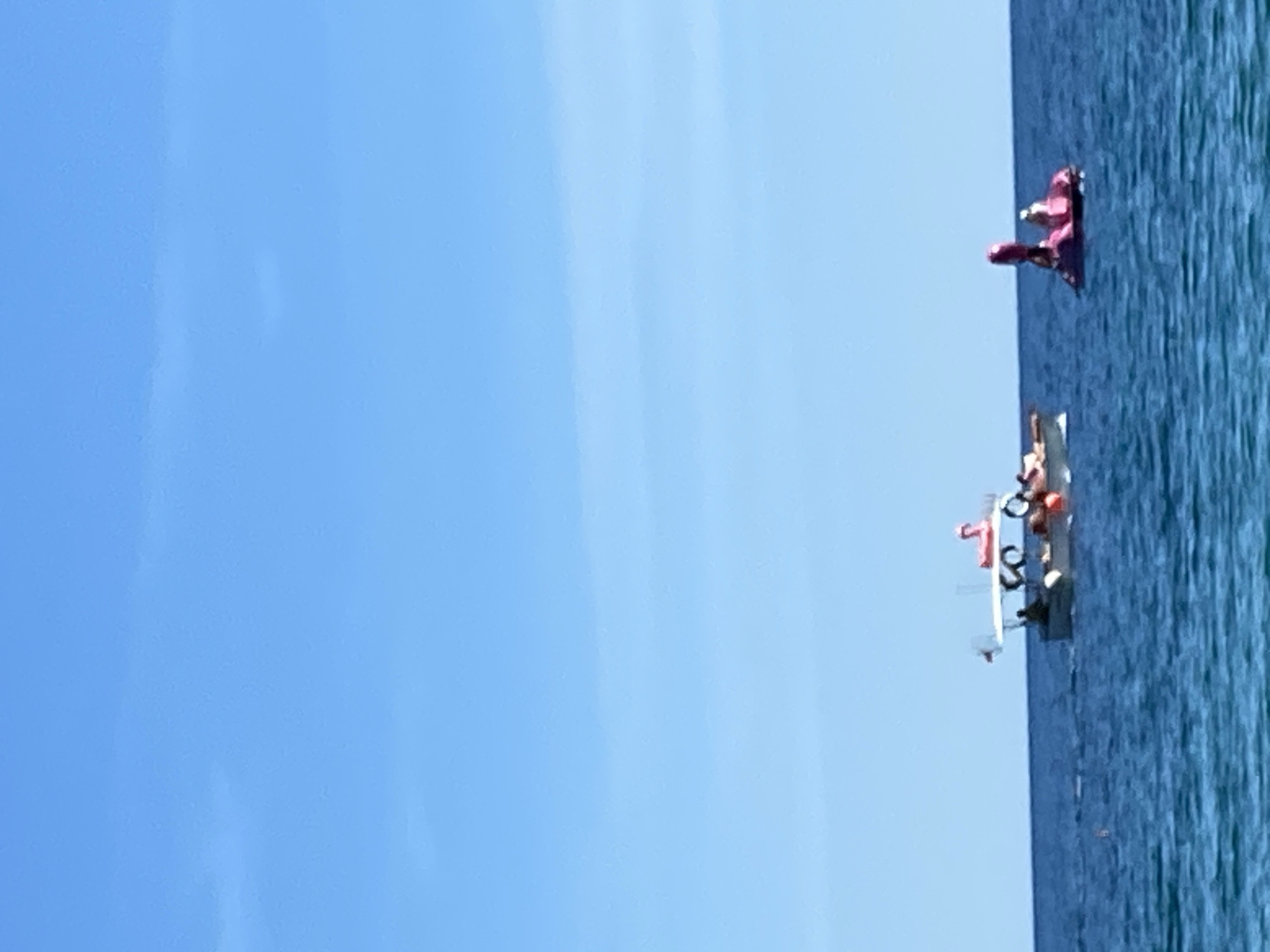 The image depicts a seascape with a clear blue sky taking up a significant portion of the frame. The sea is a deep blue color with visible surface texture and slight ripples. In the water, there is a grouping of what appear to be several small boats or rafts with people on them. There is also a pink inflatable object further out in the water. The scene is in Budva, Montenegro.