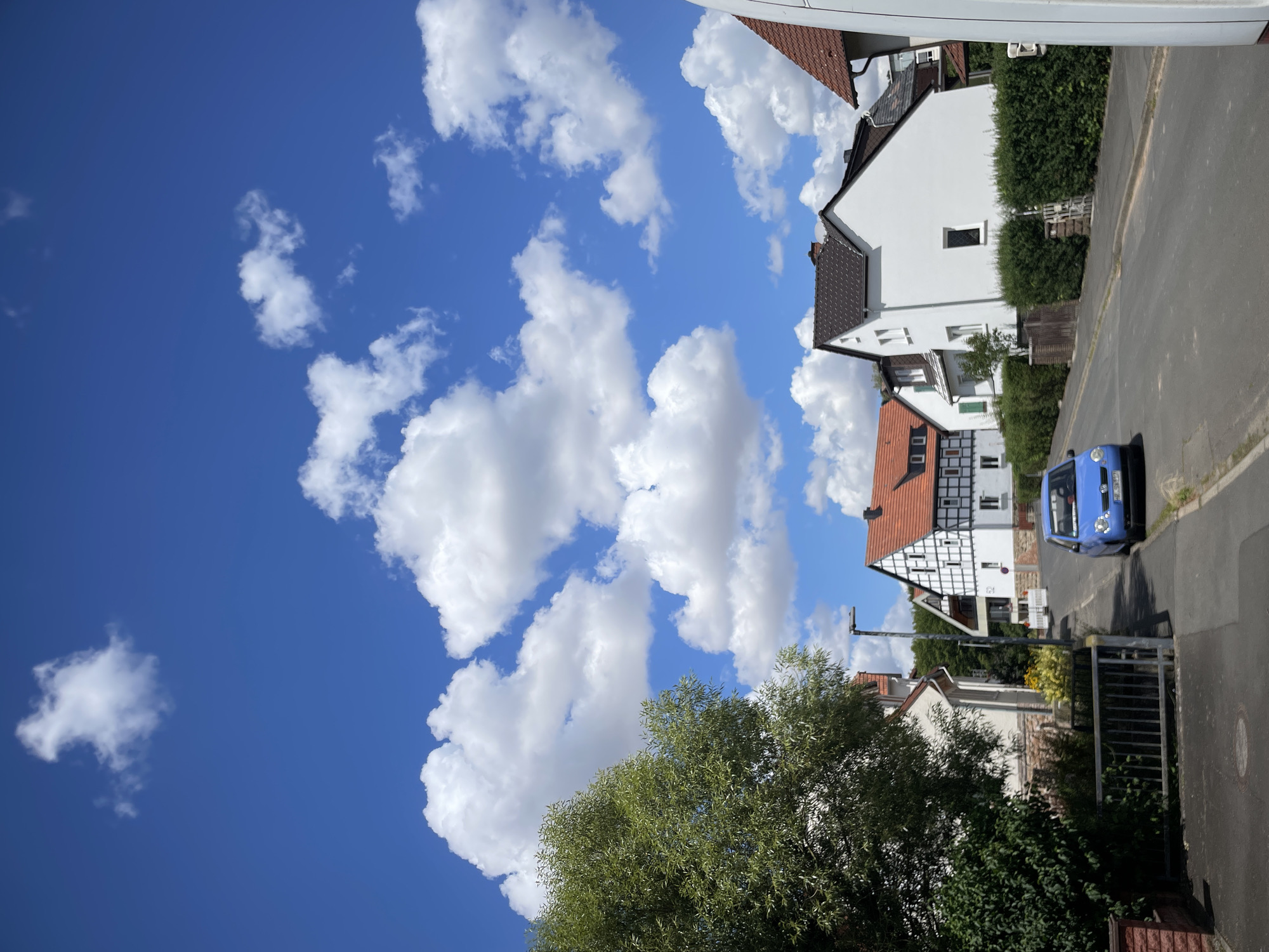 In Neukirchen, Germany, on a clear, sunny day, a residential street is depicted under a vibrant blue sky dotted with fluffy white clouds. The street curves gently to the right, bordered by a paved sidewalk and a low green hedge. On the left side of the street, a lush green tree with dense foliage stands tall. Adjacent to the tree is a house with a light-colored facade and a reddish-brown brick base. A metal fence runs along the edge of the property.
Further down the street, on the right, several houses are visible. One house features a traditional half-timbered design with white stucco walls and dark timber framing, topped with an orange-tiled roof. Next to it, a modern white house with a dark roof stands prominently. Another white house with green shutters is also present. A small blue Volkswagen Polo is parked on the right side of the street. In the background, more houses and trees are seen, suggesting a suburban neighborhood. The overall atmosphere is serene and peaceful.