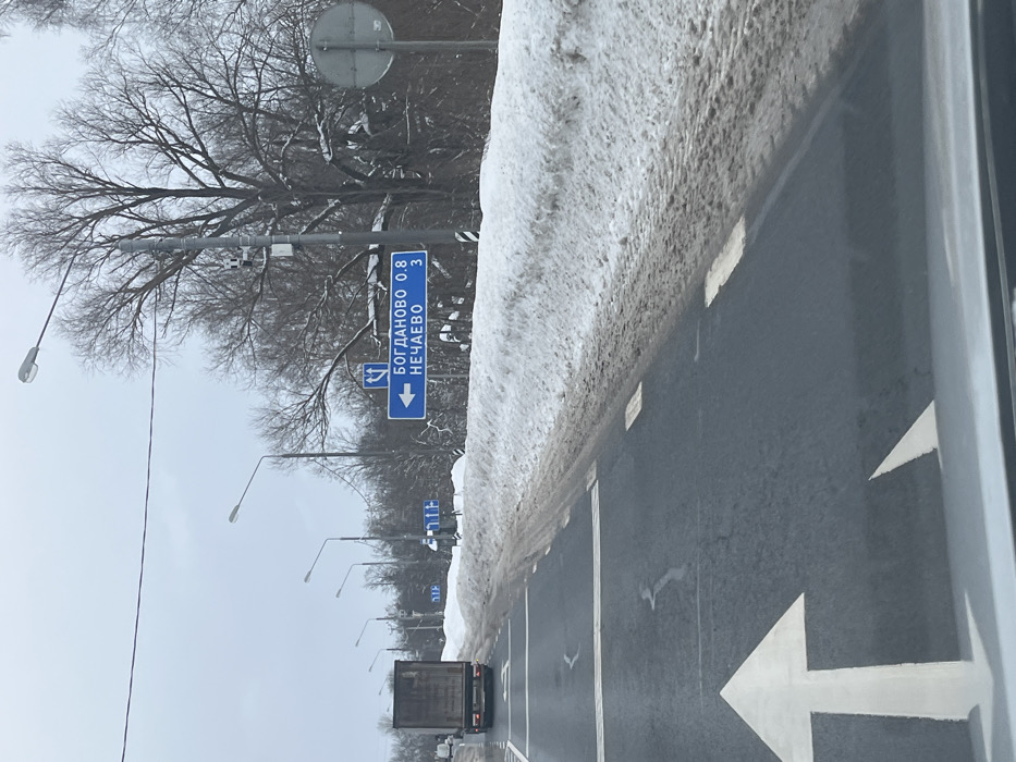 The image depicts a winter road scene from the perspective of a moving vehicle on a multi-lane asphalt road. The road surface is mostly clear, displaying white dashed and solid lane markings, and a prominent white forward-pointing arrow in the foreground. To the right, a large, soiled snow bank lines the roadside. A blue rectangular directional sign on the right, supported by a pole, indicates a left turn towards "БОГДАНОВО 0.8" (Bogdanovo, 0.8 km) and "НЕЧАЕВО 3" (Nechaevo, 3 km). A smaller white symbol on the sign illustrates a left-turn and straight-ahead option. A utility pole with a mounted camera or sensor stands near this sign. Further ahead in the left lane, a large dark-colored box truck is visible, moving away from the viewer. The background features bare deciduous trees and an overcast sky. Multiple streetlights on utility poles are visible along the road. The location is on a regional road within the vicinity of Kopylovo, Russia, based on the named localities of Bogdanovo and Nechaevo, which are situated in the Moscow Oblast. No individuals are visible.