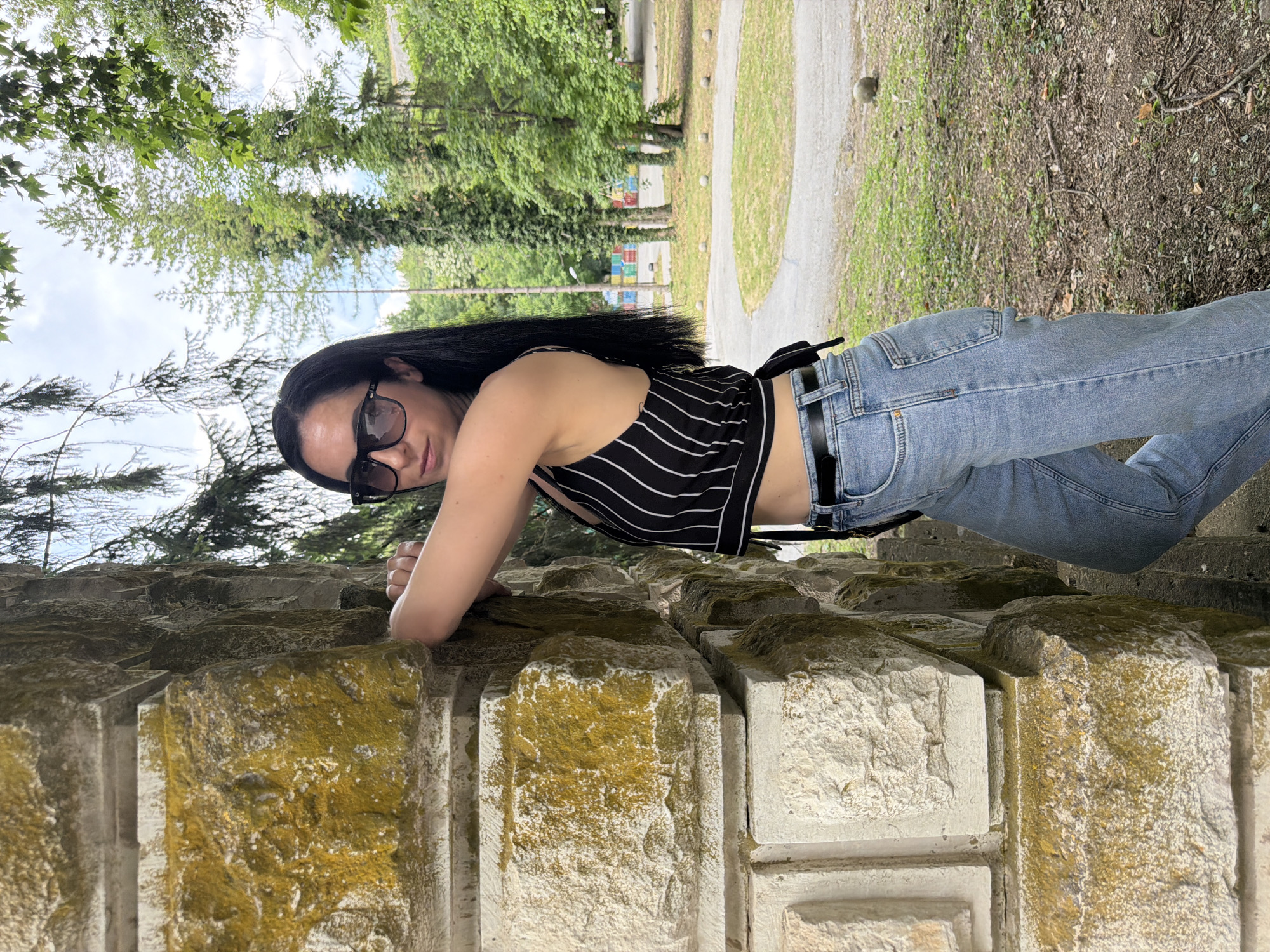 A young woman with long, dark hair, wearing sunglasses, a striped crop top, and light-wash jeans, leans against a weathered stone structure. The stone is textured and shows signs of age, with patches of moss or lichen. Behind her, a park setting is visible, with green trees and a cloudy sky. The woman's left arm is bent and resting on the stone, and her gaze is directed towards the camera. The overall scene suggests an outdoor, casual setting, likely in a park or public garden area within the city of Pleven, Bulgaria.