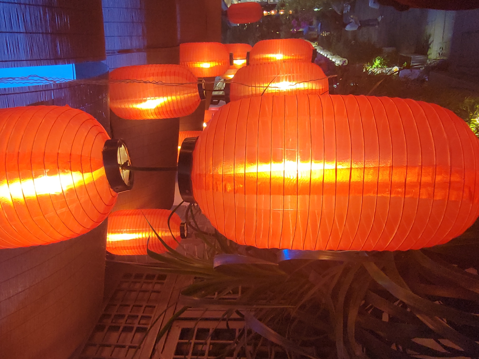 The image depicts a close-up view of a string of illuminated red lanterns, likely taken at night. The lanterns are of a traditional cylindrical shape and have a ribbed texture. The light source within each lantern is visible as a bright, concentrated glow.
In the background, other lanterns can be seen, some slightly out of focus, suggesting depth in the scene. There are hints of greenery, possibly foliage, and some structures that could be buildings or architectural features. A glimpse of what appears to be an outdoor area is visible with other lights and indistinct shapes.
The photograph was likely taken in the city of Shapingba District, China.