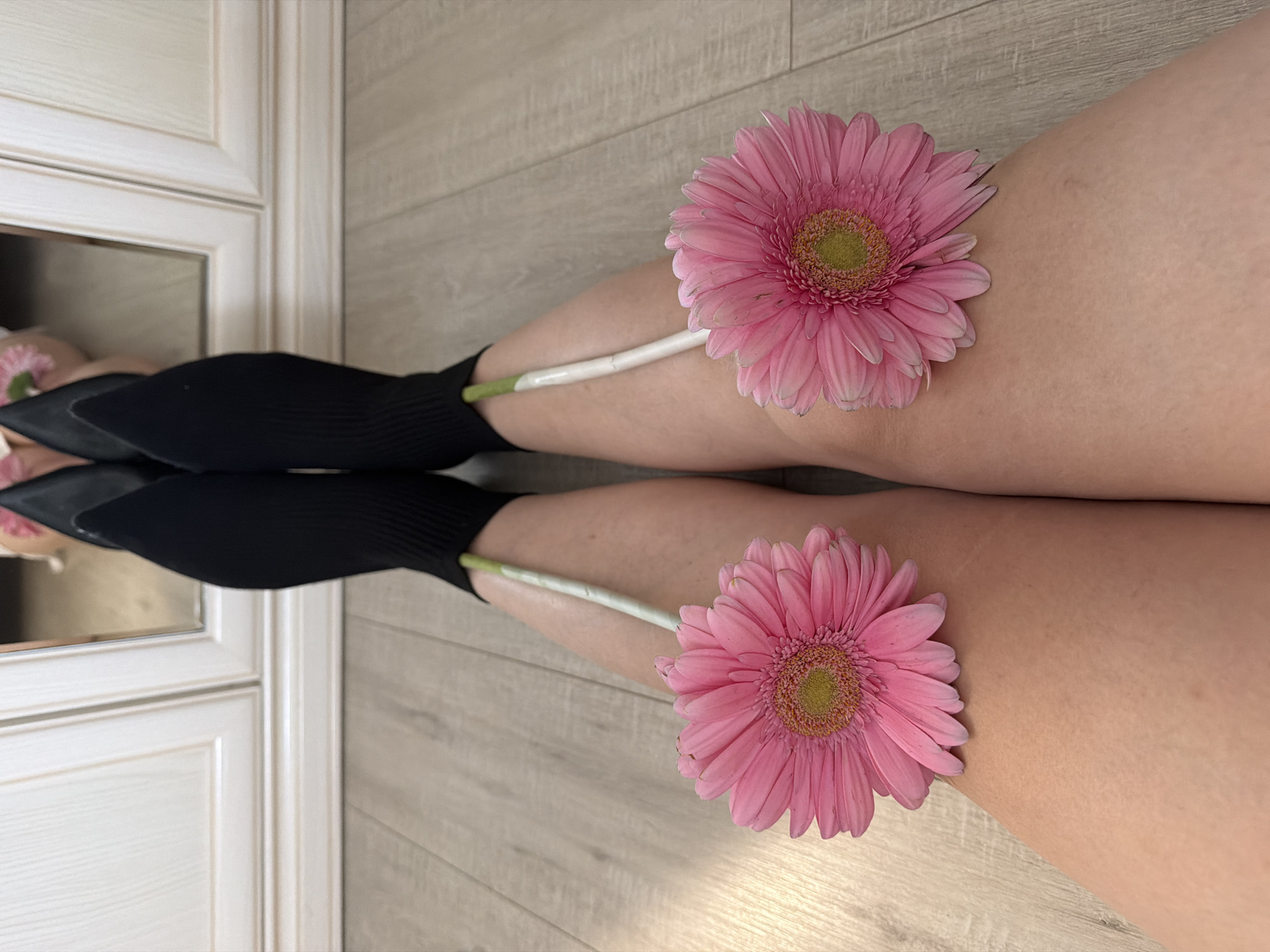 A person's legs are positioned on a light-colored wooden floor, with the lower legs covered in black sock-like shoes. Each knee is adorned with a pink gerbera daisy, with the stems extending towards the feet. A mirrored cabinet is visible in the background, reflecting the lower legs and part of the floral arrangement. The location is unknown and there is no evidence this is from Votkinsk, Russia.