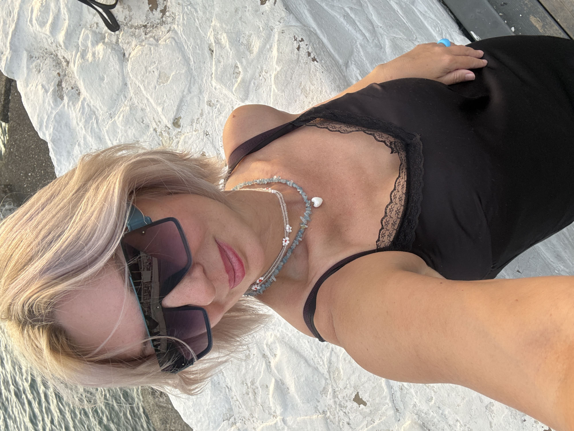 A woman is posing for a selfie outdoors, with the sea and a stone wall in the background. She has blonde hair and is wearing sunglasses, a black dress with lace trim, and necklaces. The location is a quay in Sochi, Russia.