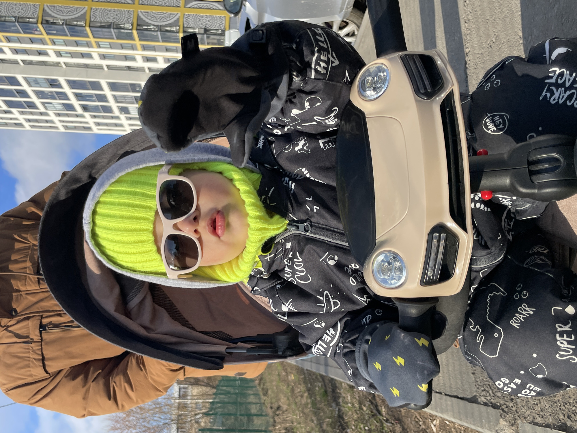 A young child is seated in a light beige ride-on toy or stroller with a car-like front and visible headlights, captured outdoors in Izhevsk, Russia, on a sunny day. The child, appearing to be a toddler or preschooler, wears a vibrant neon green knitted beanie, white-framed sunglasses, a black jacket adorned with white patterns and text, and black mittens featuring yellow lightning bolts. The child is making a playful "duck face" or pouting expression towards the viewer.
Part of an adult's arm, dressed in a brown jacket, is visible on the left, suggesting they are accompanying or pushing the child. The child's clothing is covered in various phrases and cartoon-like graphics. Visible text includes "HELLO", "WILD", "COOL", "SUPE", "EASY GO", "ROARR", "SUPER", "SCARY FACE", and "HEY DUDE".
The background features a clear blue sky and a tall, modern building with numerous square windows framed in yellow, indicating an urban setting. The ground is paved, resembling a sidewalk or pedestrian area, with some sparse greenery or trees further in the distance. The scene depicts a bright, cheerful daytime outing.