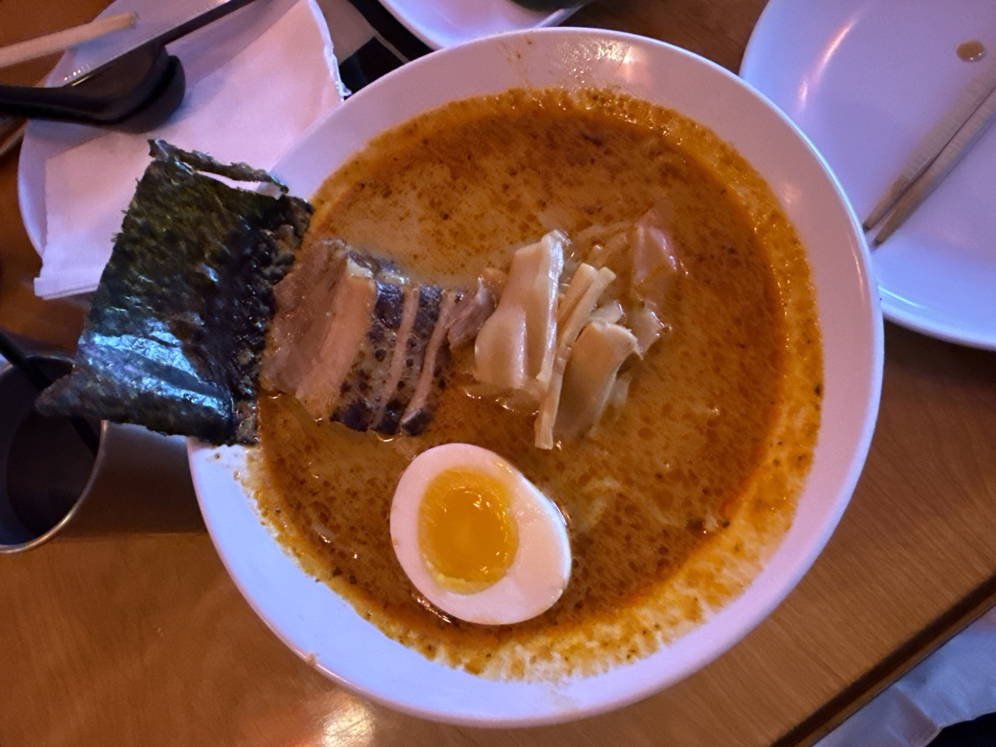 A close-up, overhead shot features a bowl of ramen placed on a wooden table. The ramen contains broth with orange and brown hues, a sliced soft-boiled egg, sliced pork belly with visible sear marks, bamboo shoots, and a piece of dried seaweed (nori). To the left of the bowl, there are eating utensils. In the background, partially visible are plates. The location is estimated to be in Hollywood, United States of America.