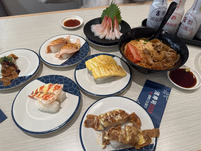 The scene depicts a dining table laden with various Japanese food items at a Hama Sushi restaurant, specifically identified as the Hama Sushi Raffles City Chongqing store in Yuzhong District, China. The table surface is light-colored.
**Food Items:**
- A black ceramic bowl contains a noodle dish with a reddish-brown broth, topped with sliced beef, caramelized onions, and a wedge of red tomato. A black spoon is partially submerged in the bowl.
- A small, dark-colored rectangular plate holds several peeled, tail-on raw shrimp, accompanied by a single green shiso leaf.
- Five round white ceramic plates, each adorned with a blue wave or geometric pattern along the rim, display various sushi and nigiri:
- One plate contains two pieces of seared salmon nigiri.
- Another plate holds two pieces of tamagoyaki (Japanese omelet) nigiri.
- A third plate features two pieces of nigiri topped with red-orange shellfish, dressed with a yellowish sauce.
- A fourth plate presents two pieces of nigiri topped with grilled or seared meat and a light sauce drizzle.
- A fifth plate displays two pieces of nigiri with dark, glazed toppings, garnished with green herbs.
- Two small white rectangular dishes contain dark brown liquid, identified as soy sauce. One dish also contains a small amount of green wasabi paste.
**Condiments and Accessories:**
- A black rectangular tray holds three white plastic squeeze bottles with Japanese and Chinese labels, indicating different types of soy sauce, including reduced salt and brewed varieties.
- A blue rectangular packet of "Hama Sushi" brand 75% reduced salt soy sauce is positioned on the table.
**Environment and Activity:**
The arrangement of dishes suggests a meal either just served or in progress for at least one, likely multiple, diners. The background is softly blurred, showing light-colored seating upholstery consistent with an indoor restaurant setting. No individuals are visible.